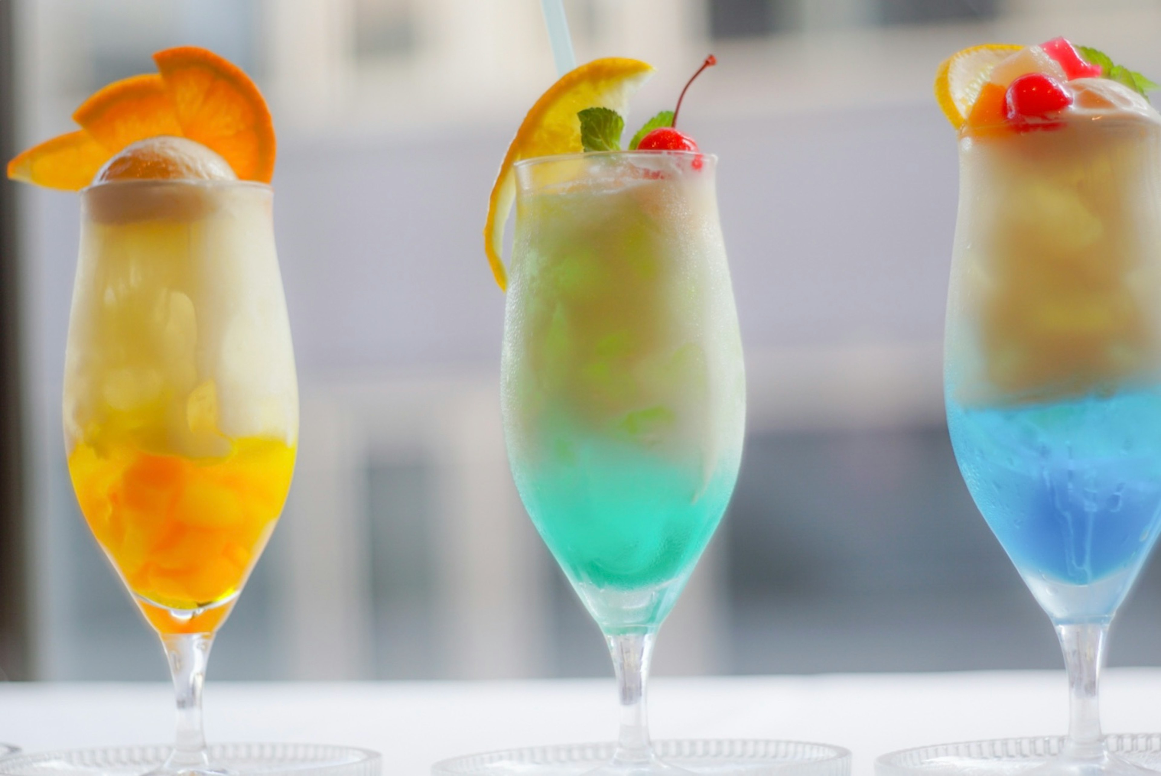 Farbige Darstellung von drei Cocktails mit blauen, gelben und grünen Schichten garniert mit Orange und Kirsche