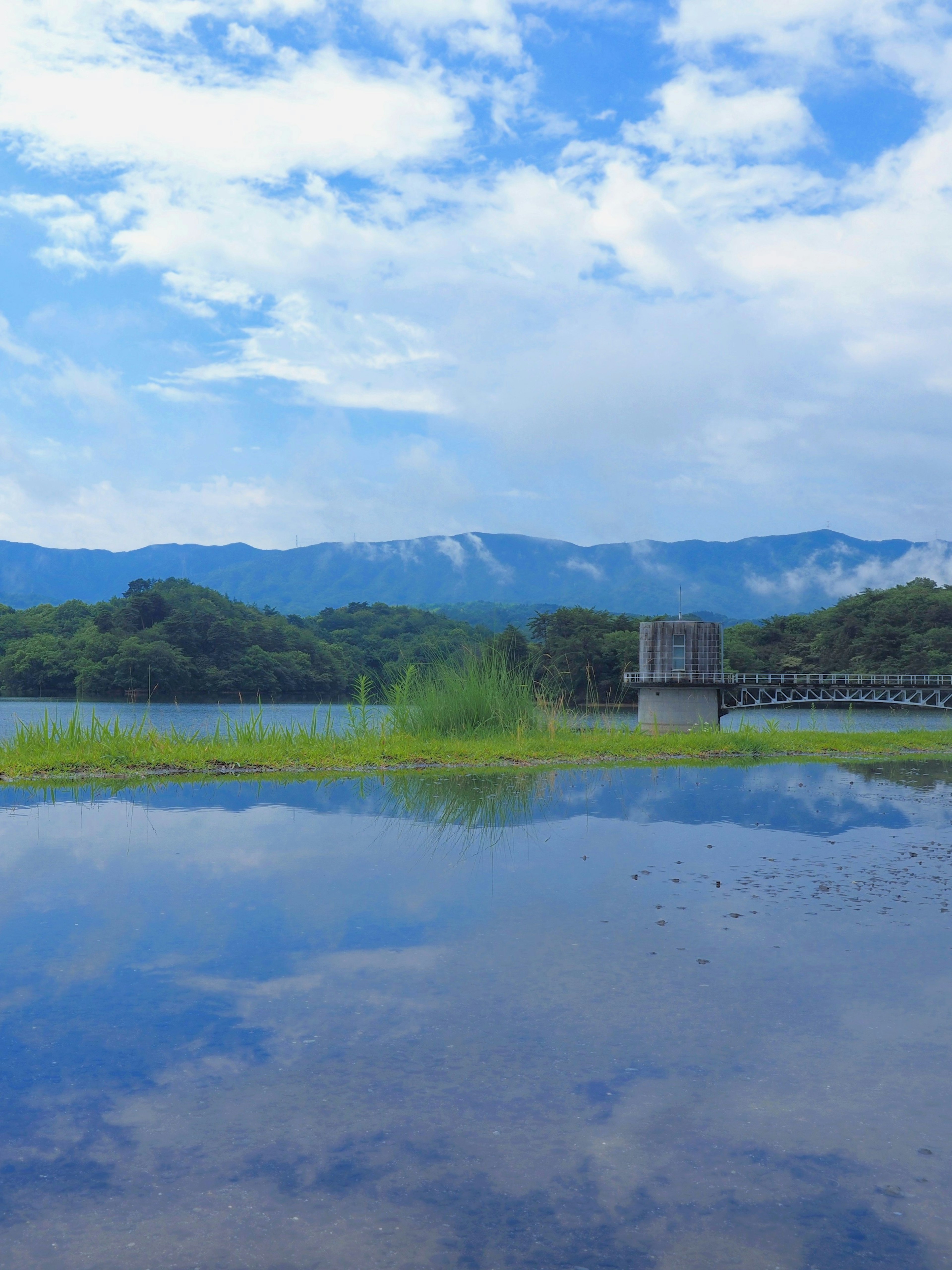 Pemandangan danau yang mencerminkan langit biru dan awan dengan pegunungan di latar belakang