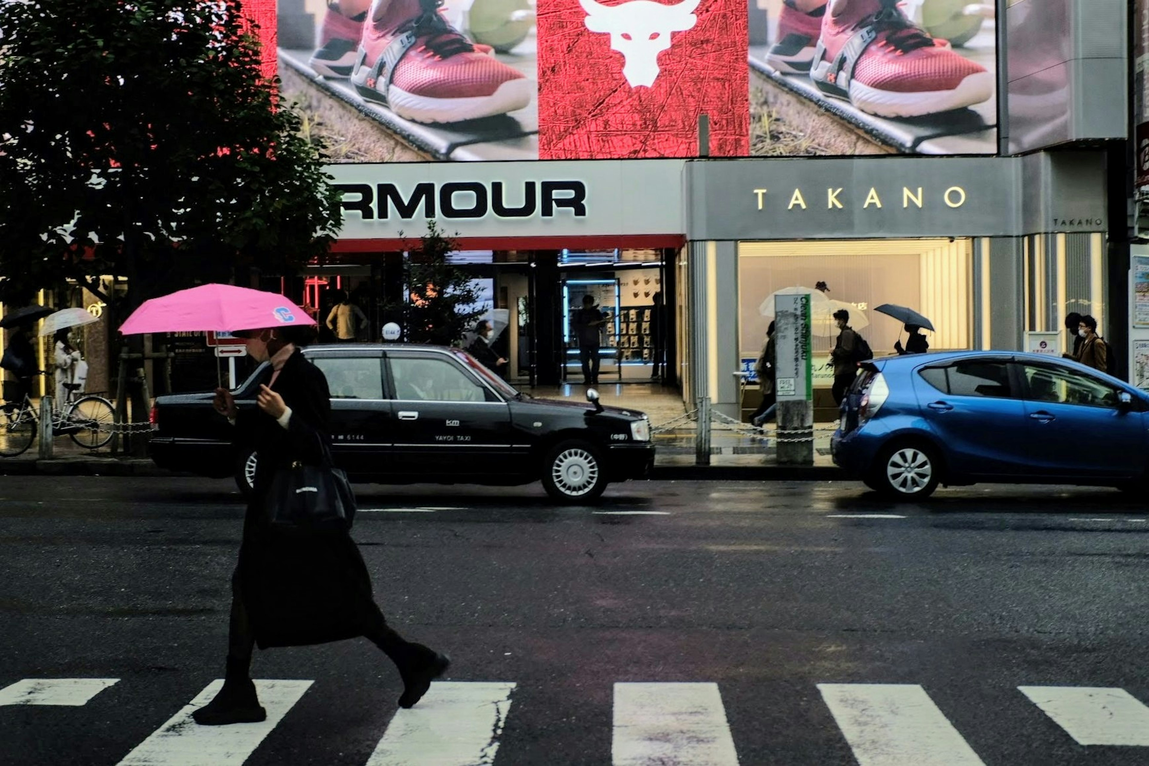 雨の中でピンクの傘を持って横断歩道を渡る人とショップの看板