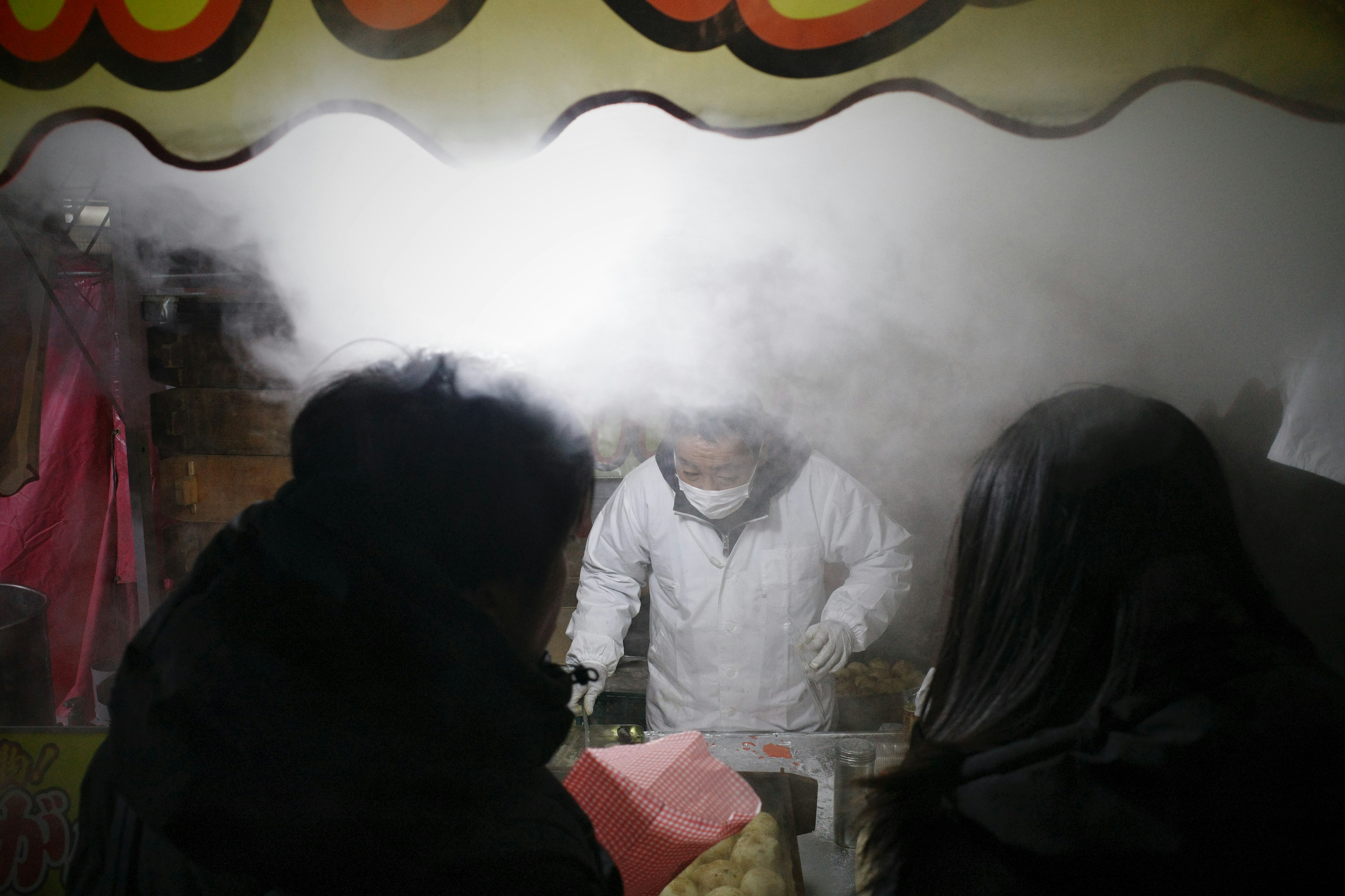 Vendor in white preparing food at a smoky stall with two customers