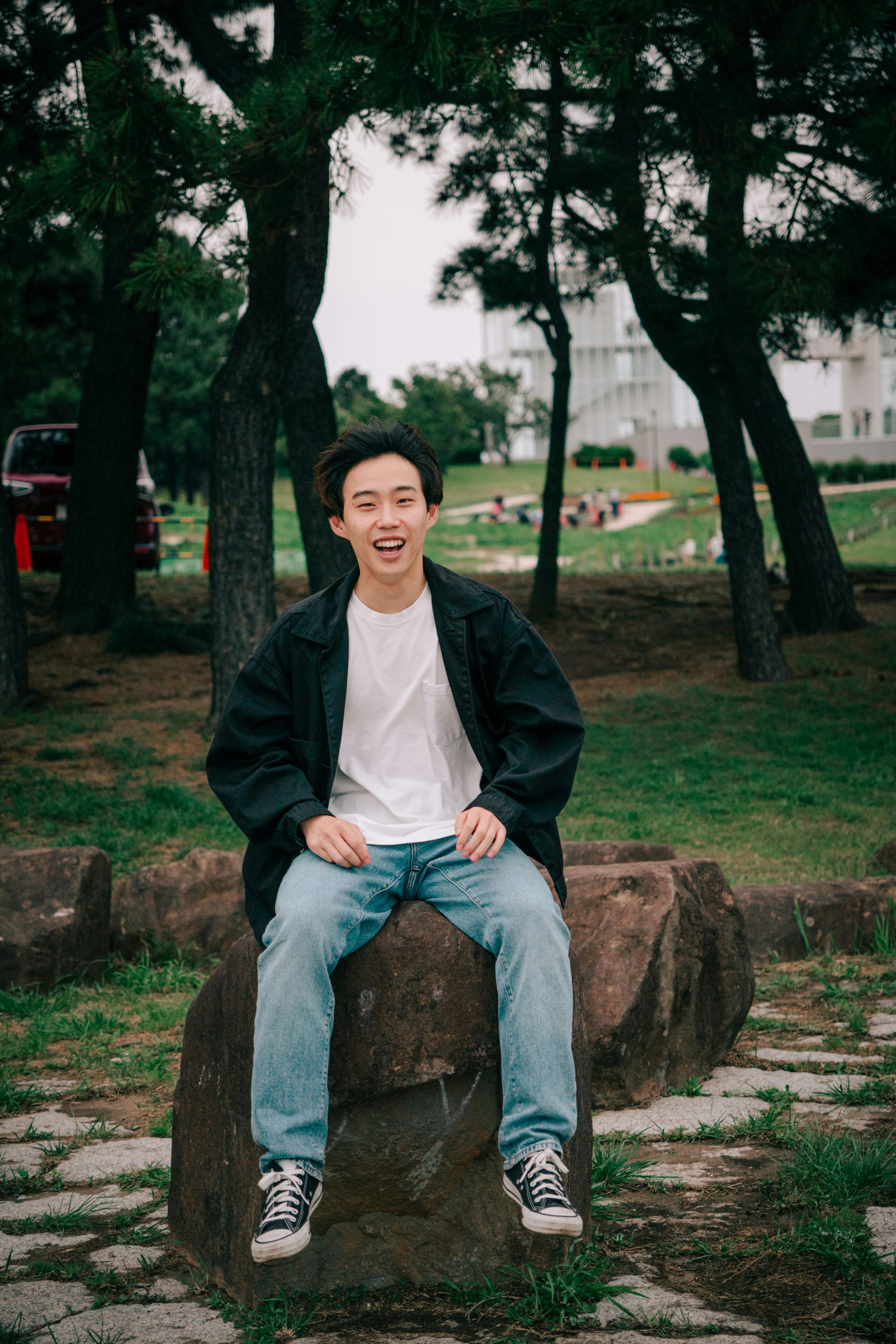 Joven sentado en una piedra en un parque sonriendo árboles y un edificio al fondo