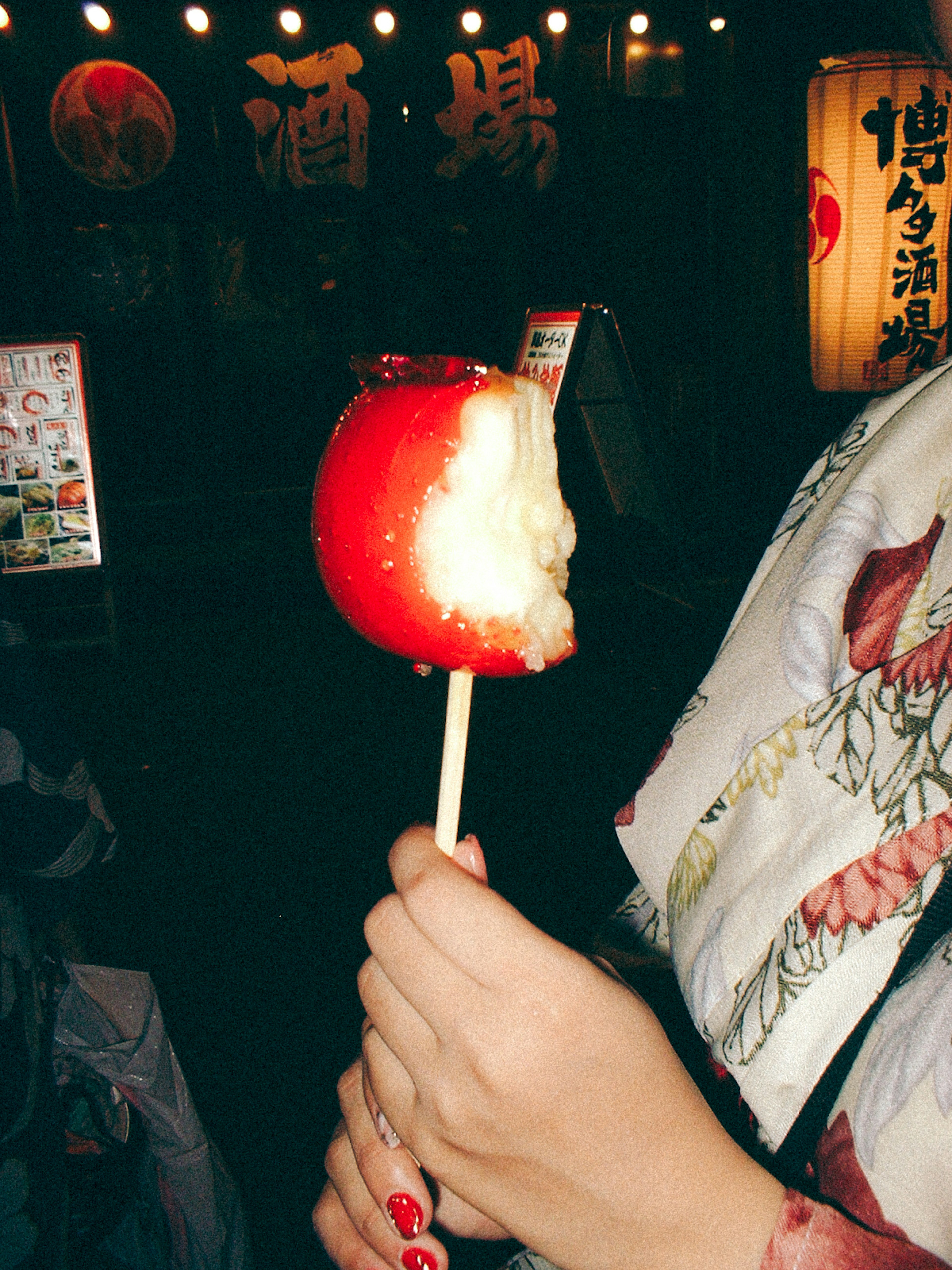 A woman's hand holding a red candy apple with a bite taken out and lanterns in the background