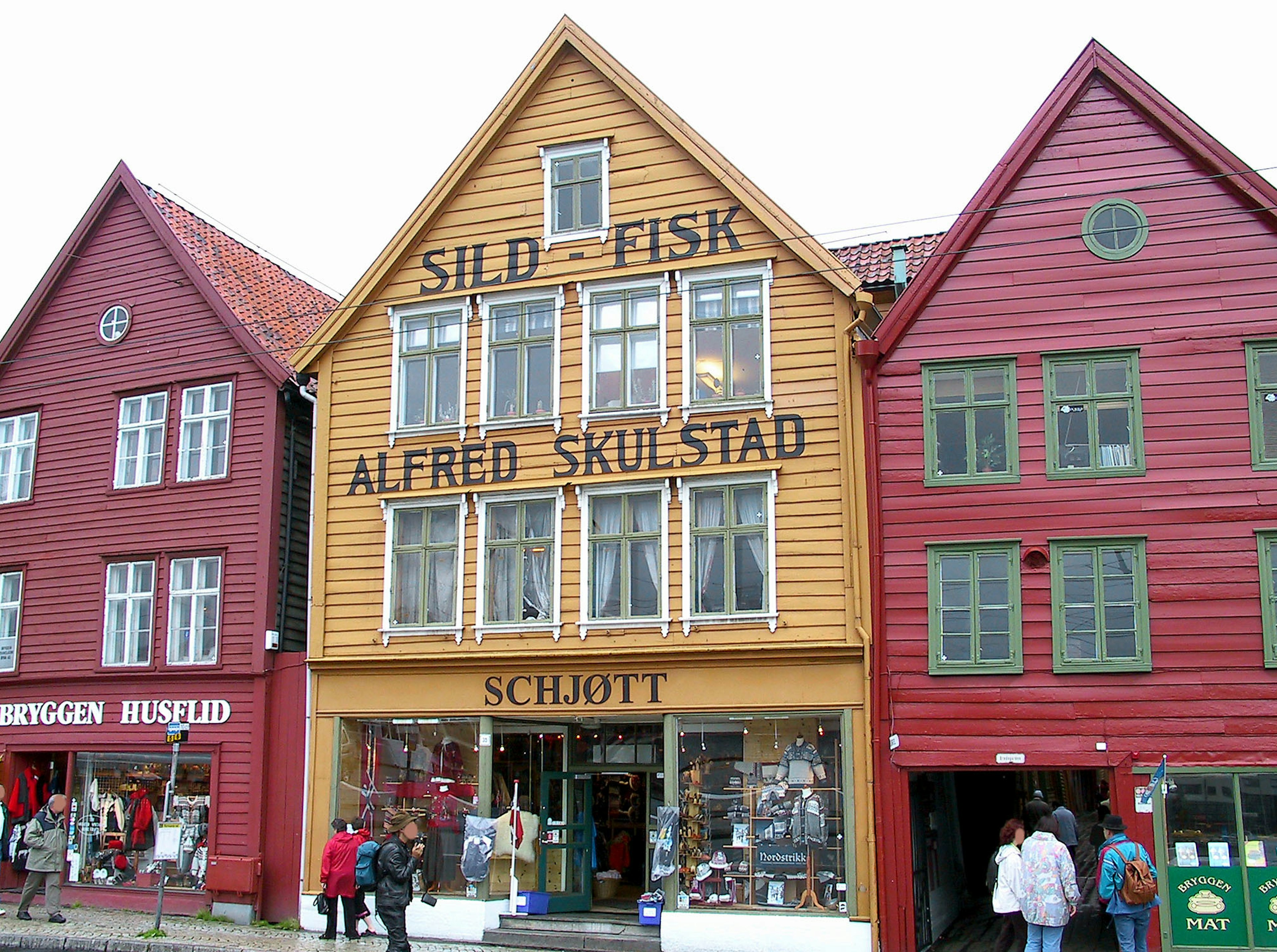 Bunte Holzbauten säumen die Straße mit einem gelben Gebäude, das die Schilder SILD-FISK und ALFRED SKULSTAD zeigt