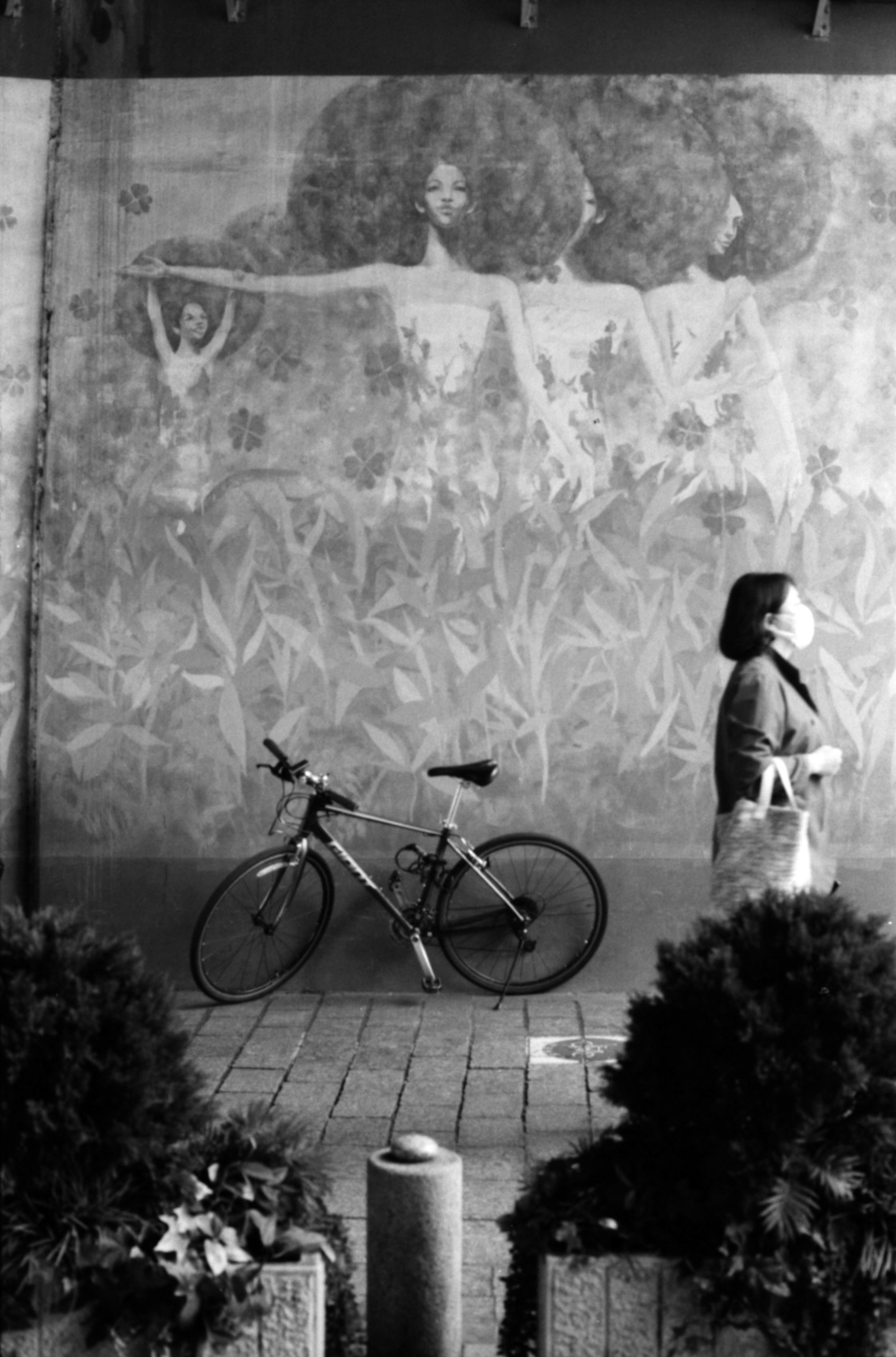 Black and white photo of a woman walking in front of a mural with a bicycle