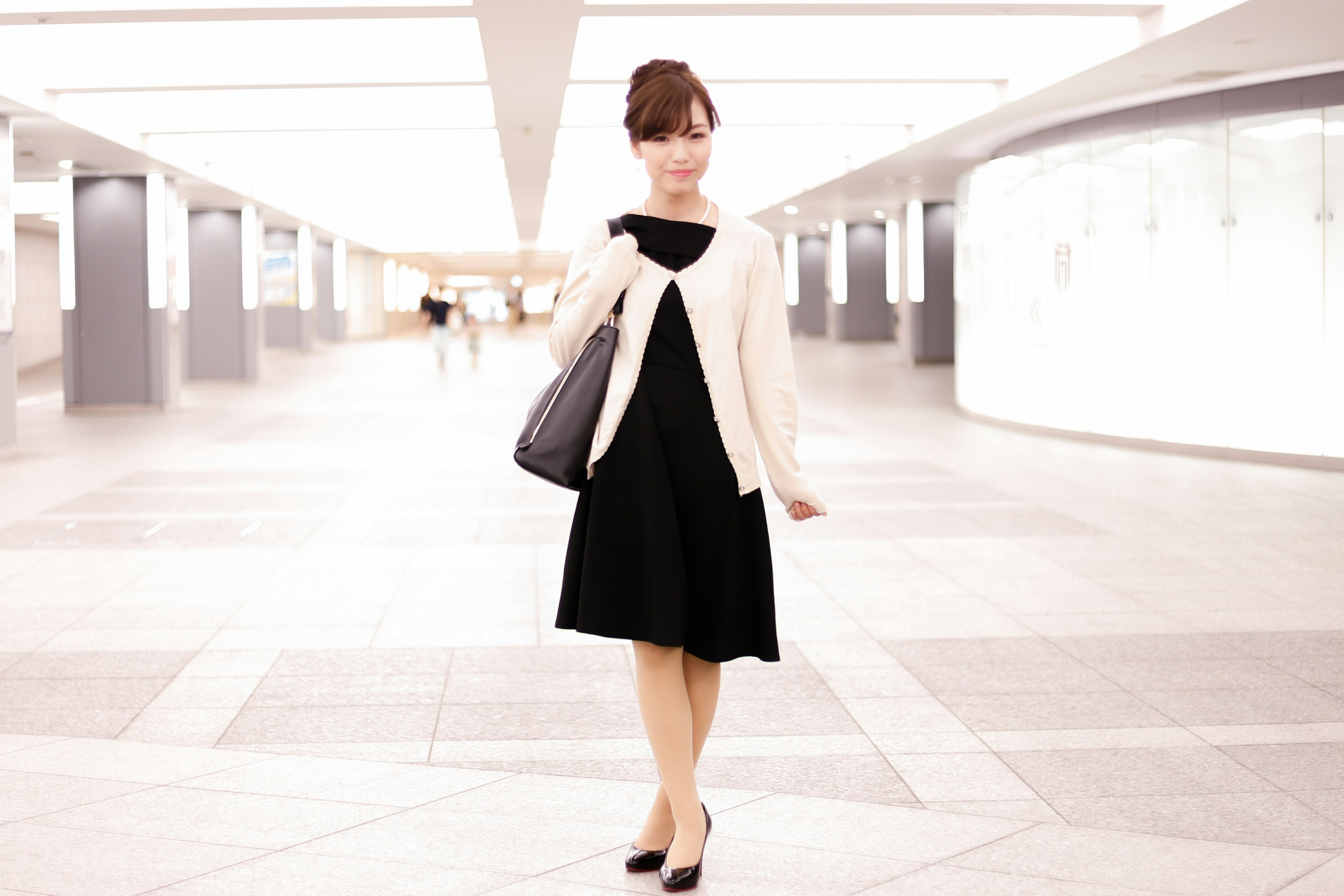 A woman in a black dress wearing a white cardigan stands elegantly in a well-lit space