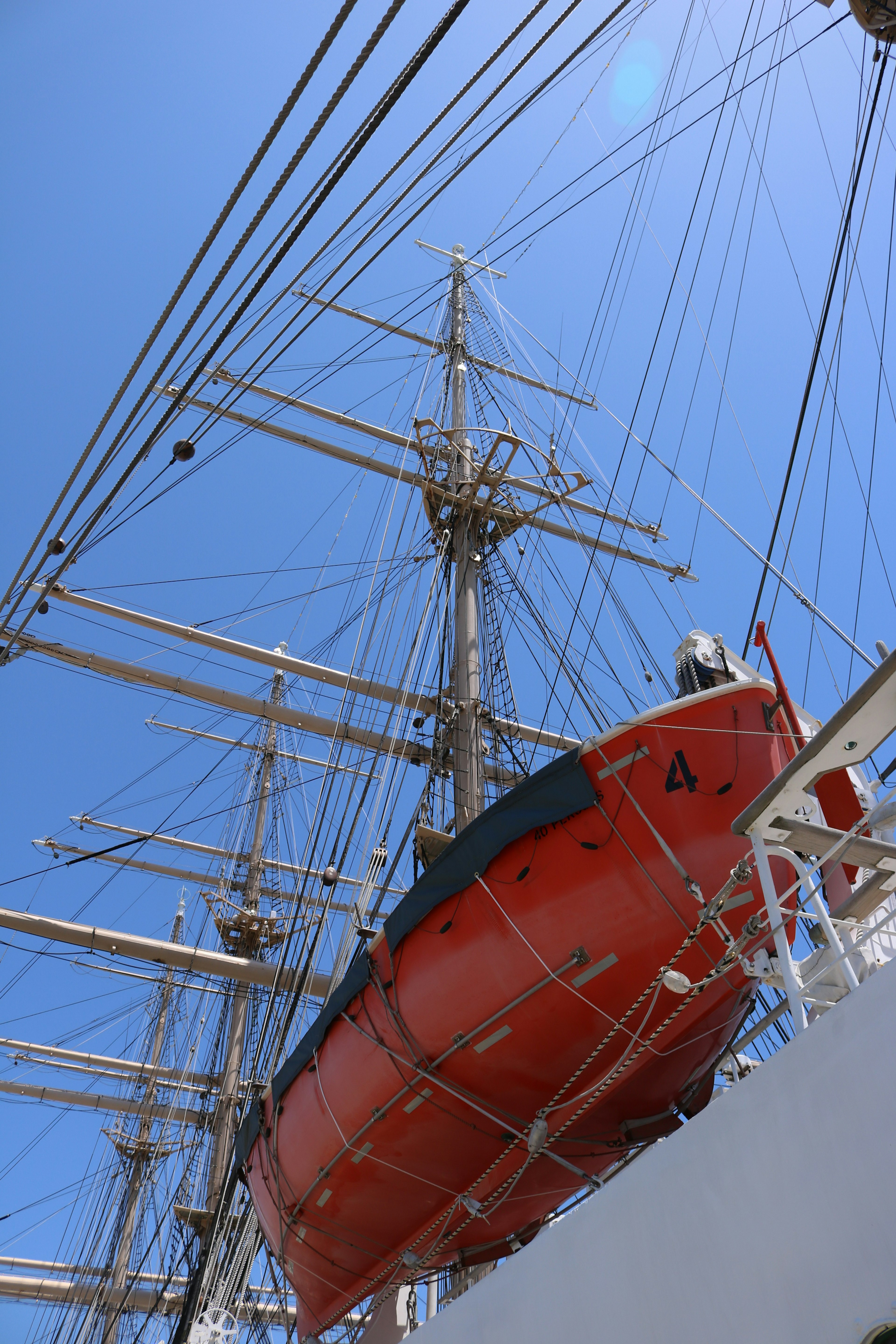 Canot de sauvetage orange suspendu haut sur un grand voilier sous un ciel bleu clair