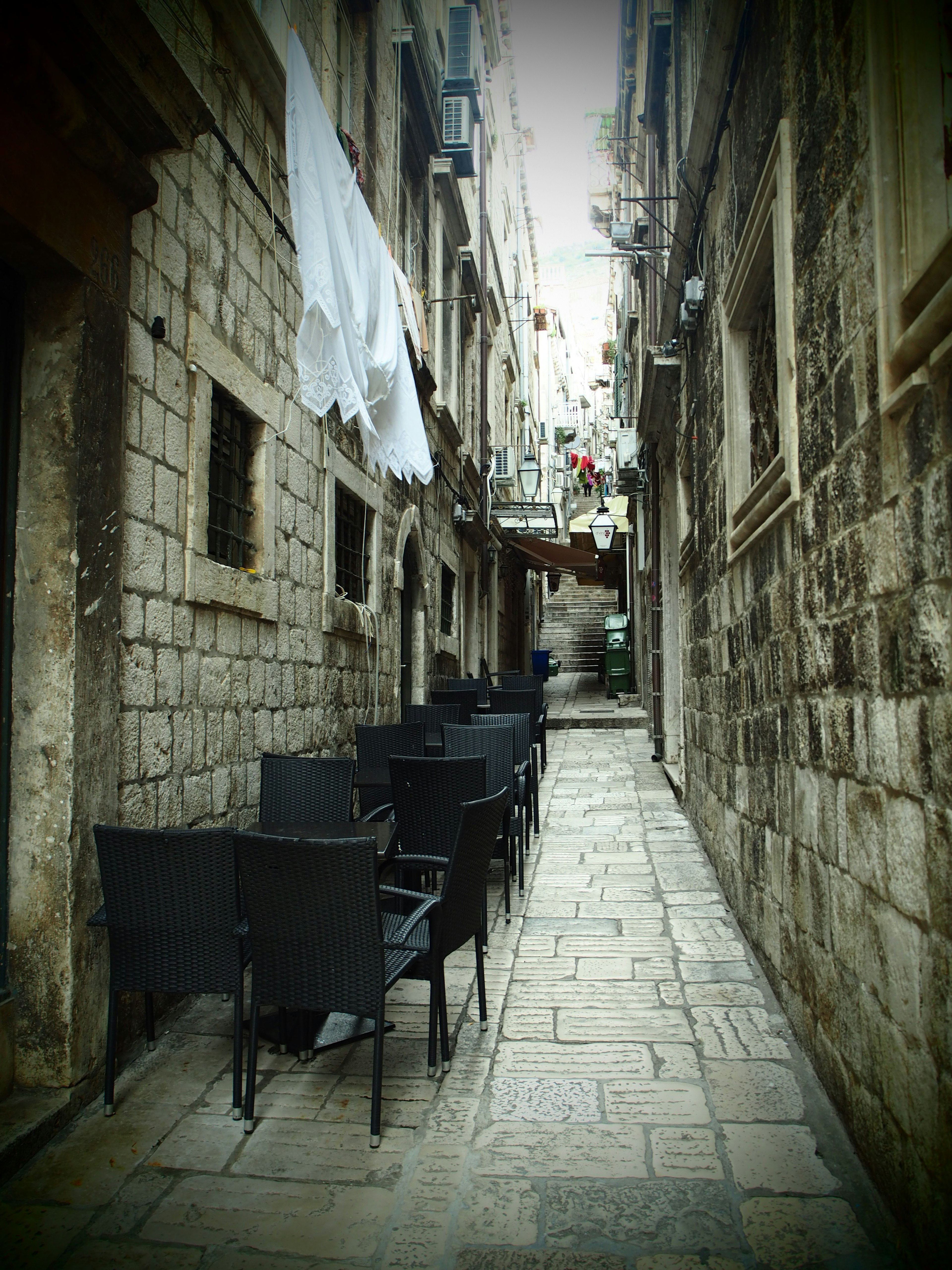 Callejón estrecho de piedra con sillas negras alineadas y ropa blanca colgando