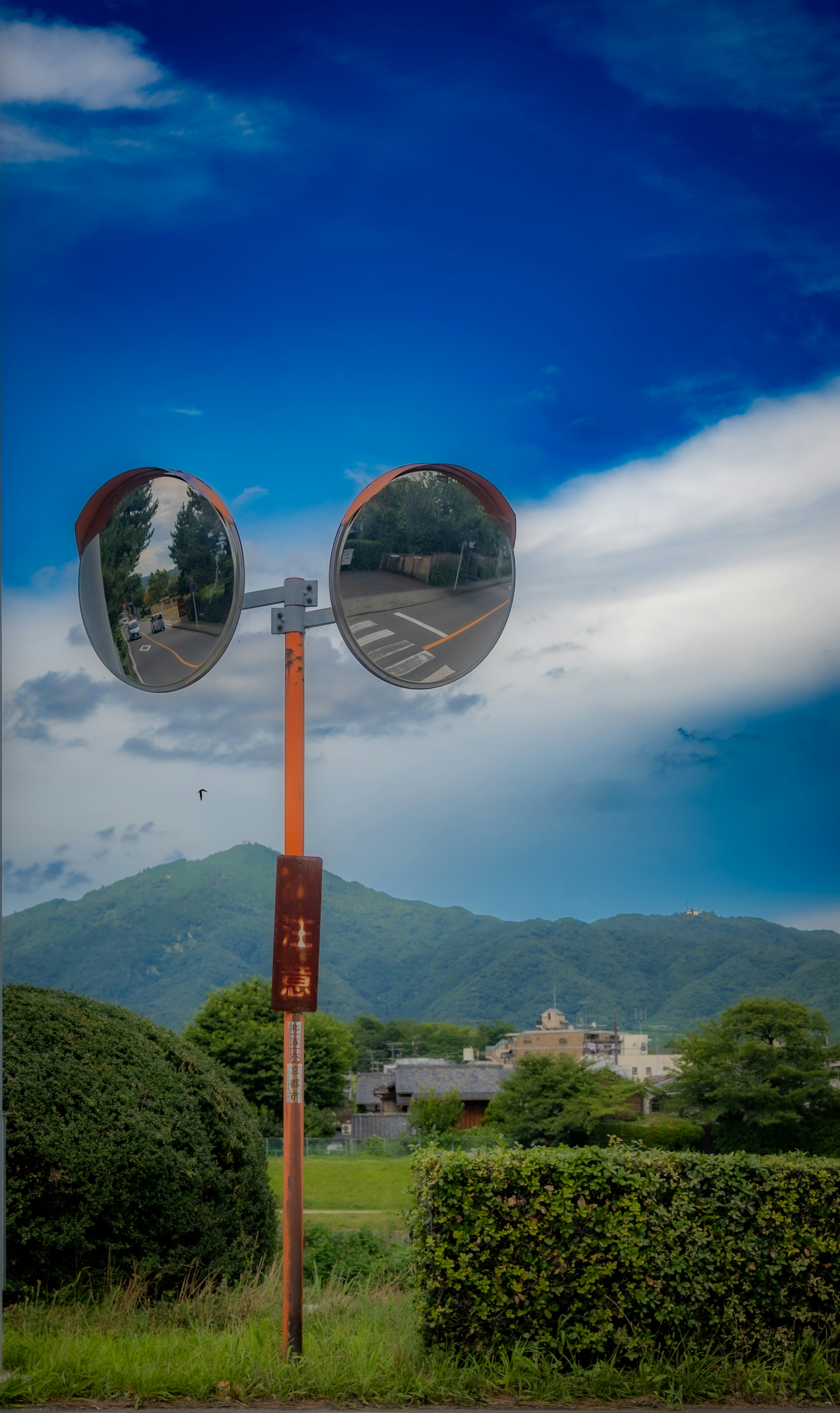 Large convex mirrors mounted on a pole under a blue sky with mountains and buildings in the background