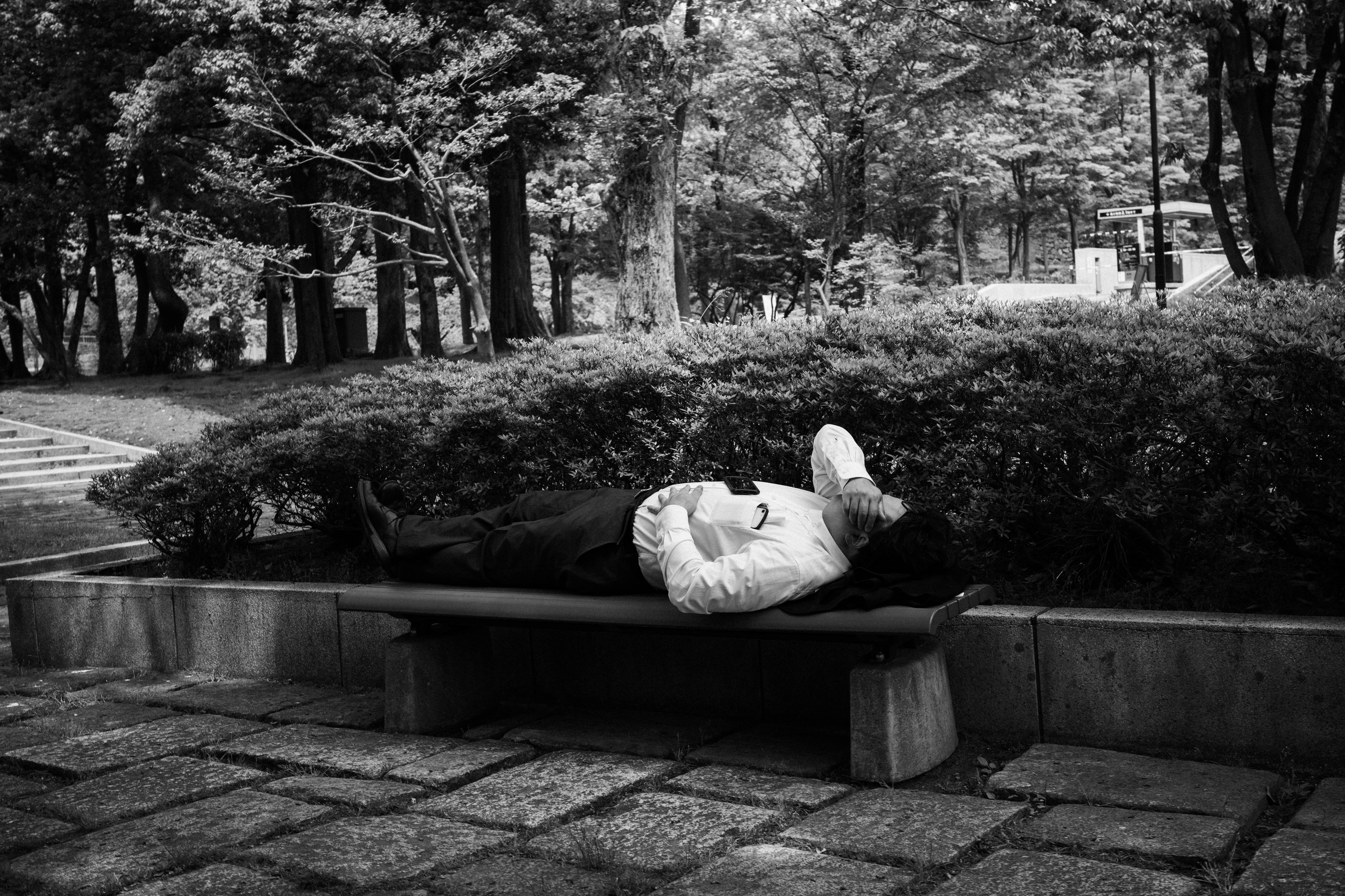 A man lying on a bench in a park surrounded by green shrubs black and white photo