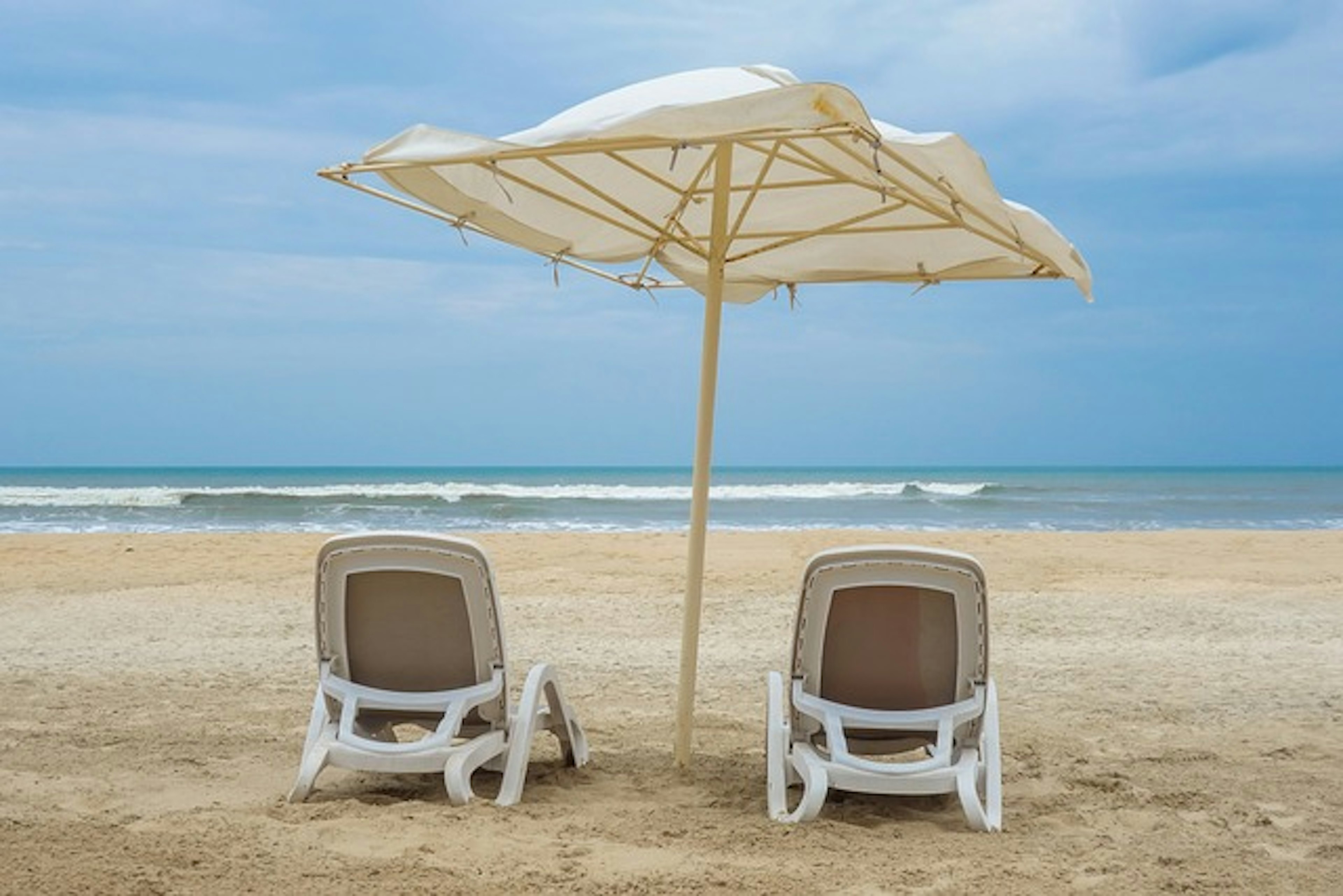 Zwei weiße Liegestühle unter einem Sonnenschirm am Strand