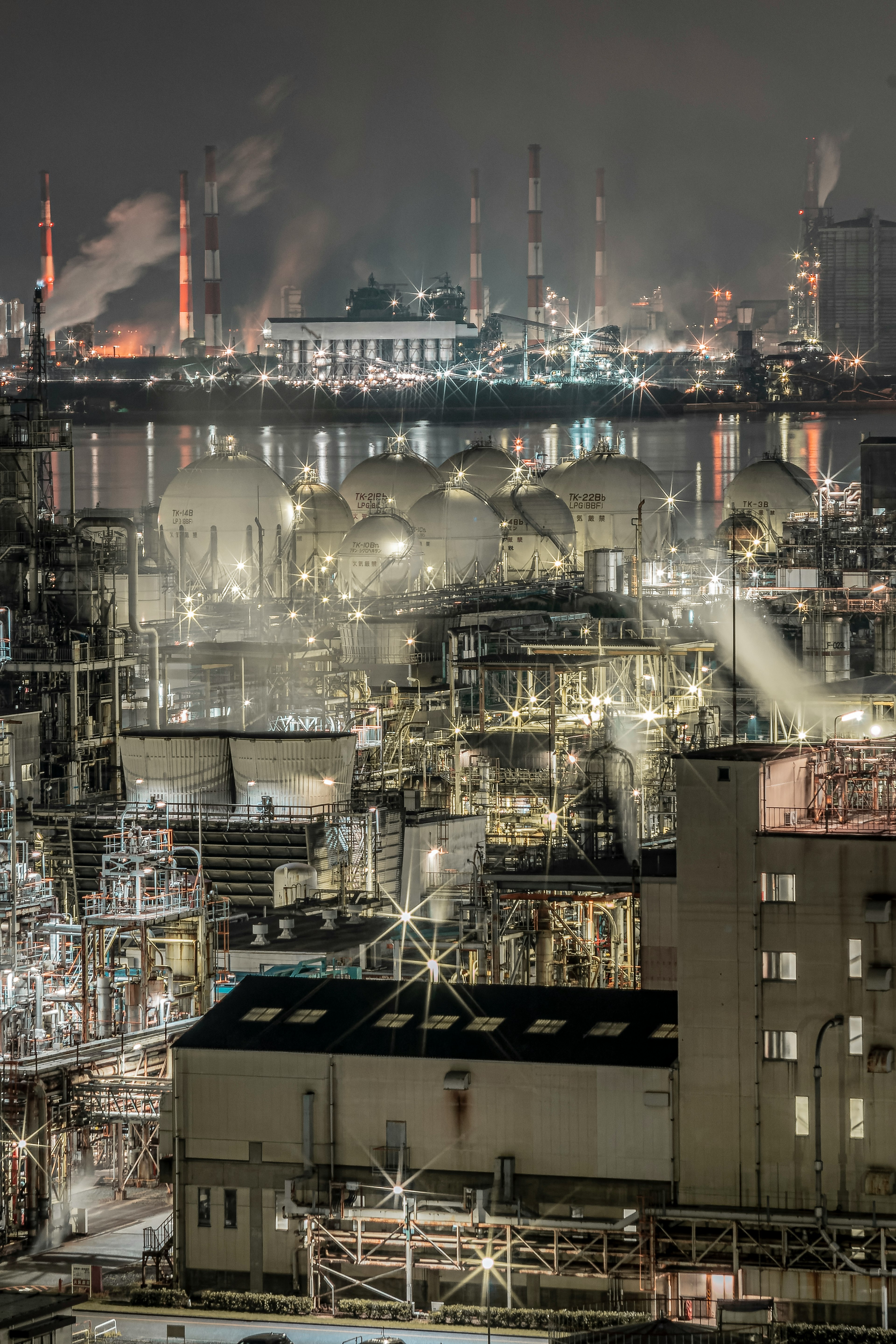 Industrial landscape at night featuring tanks and smokestacks with smoke and lights