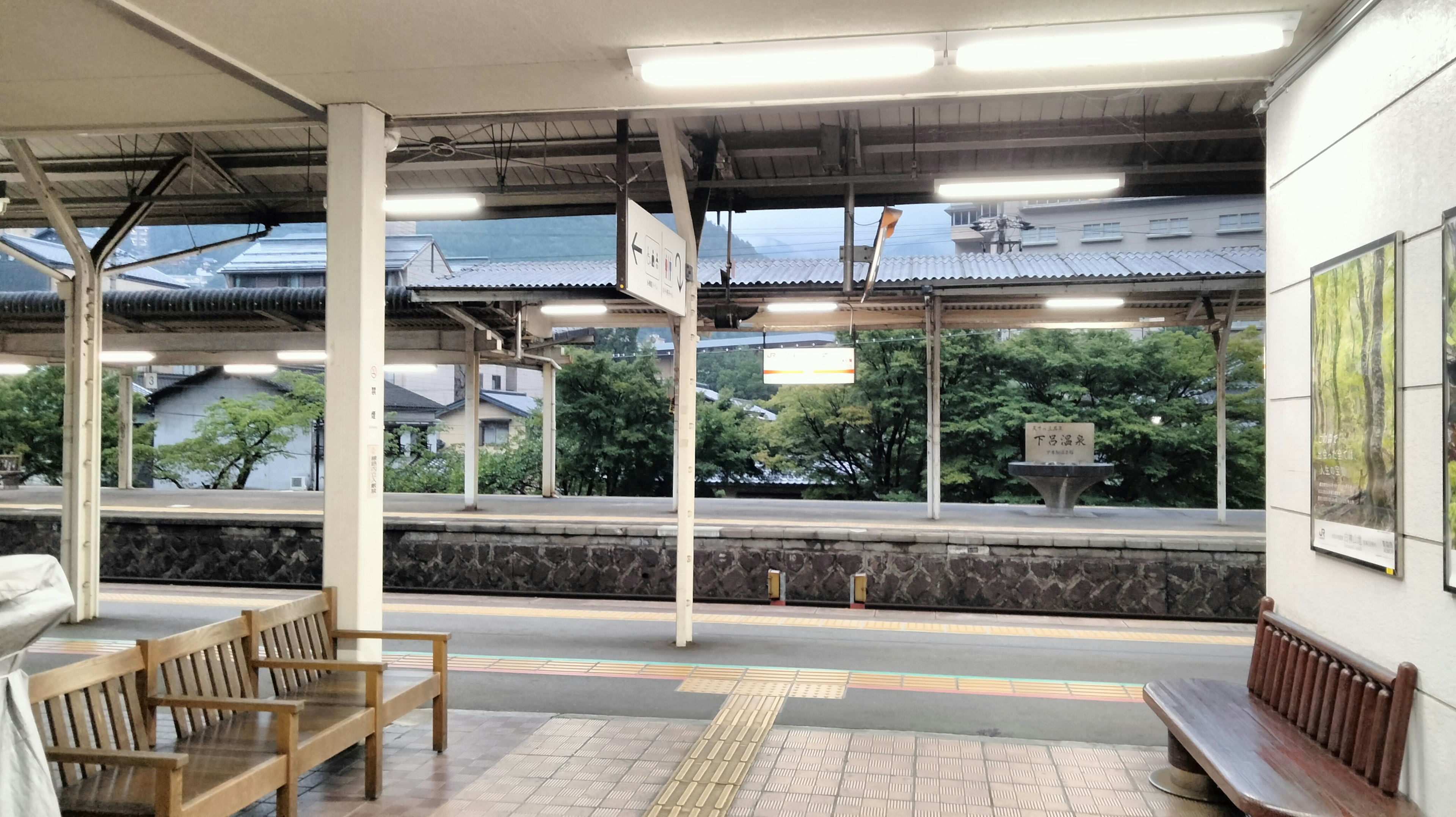 View of a train station platform with benches and overhead lights featuring a green landscape