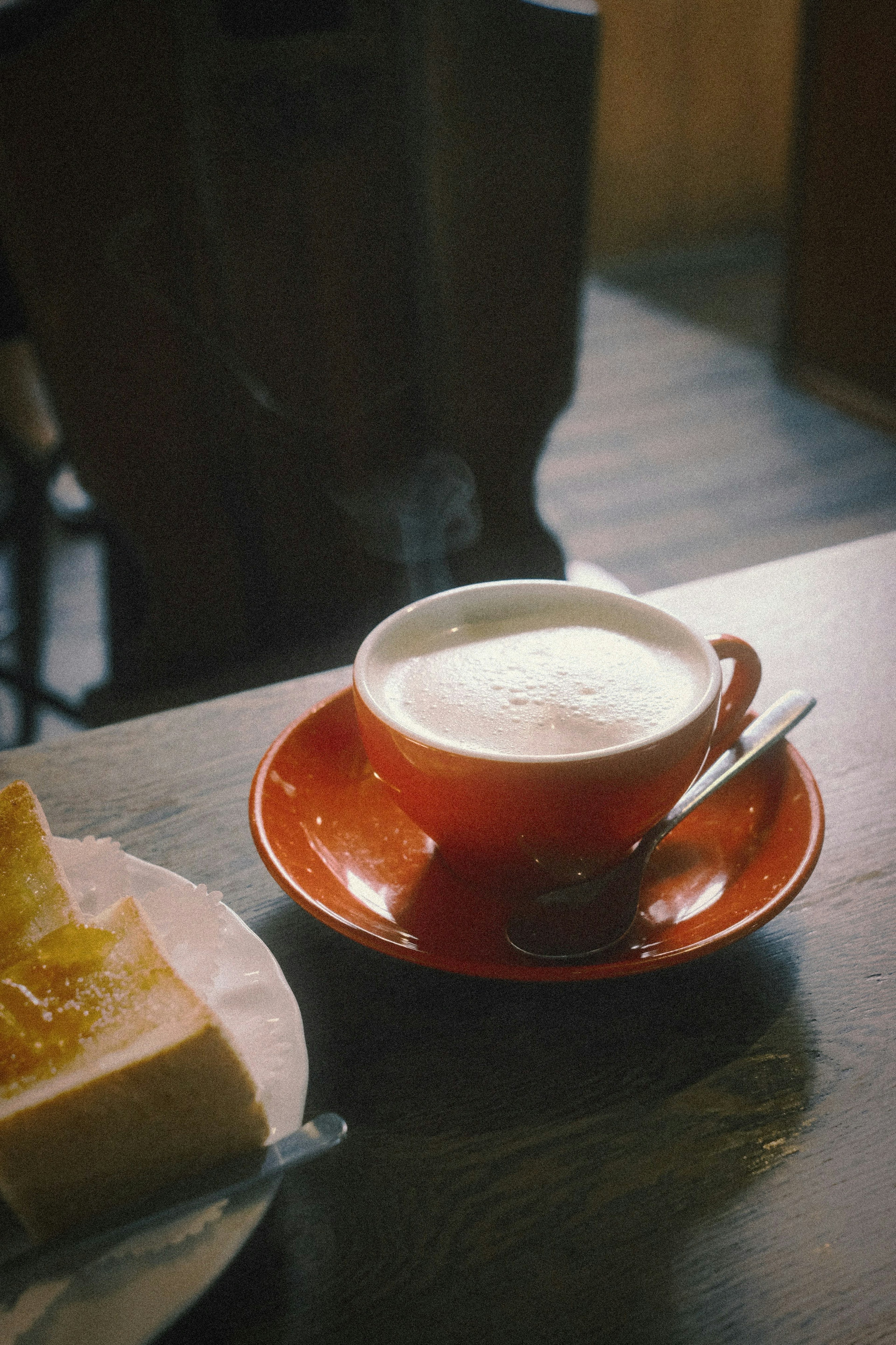 Dampfende Milch in einer roten Tasse mit einem Stück Kuchen daneben