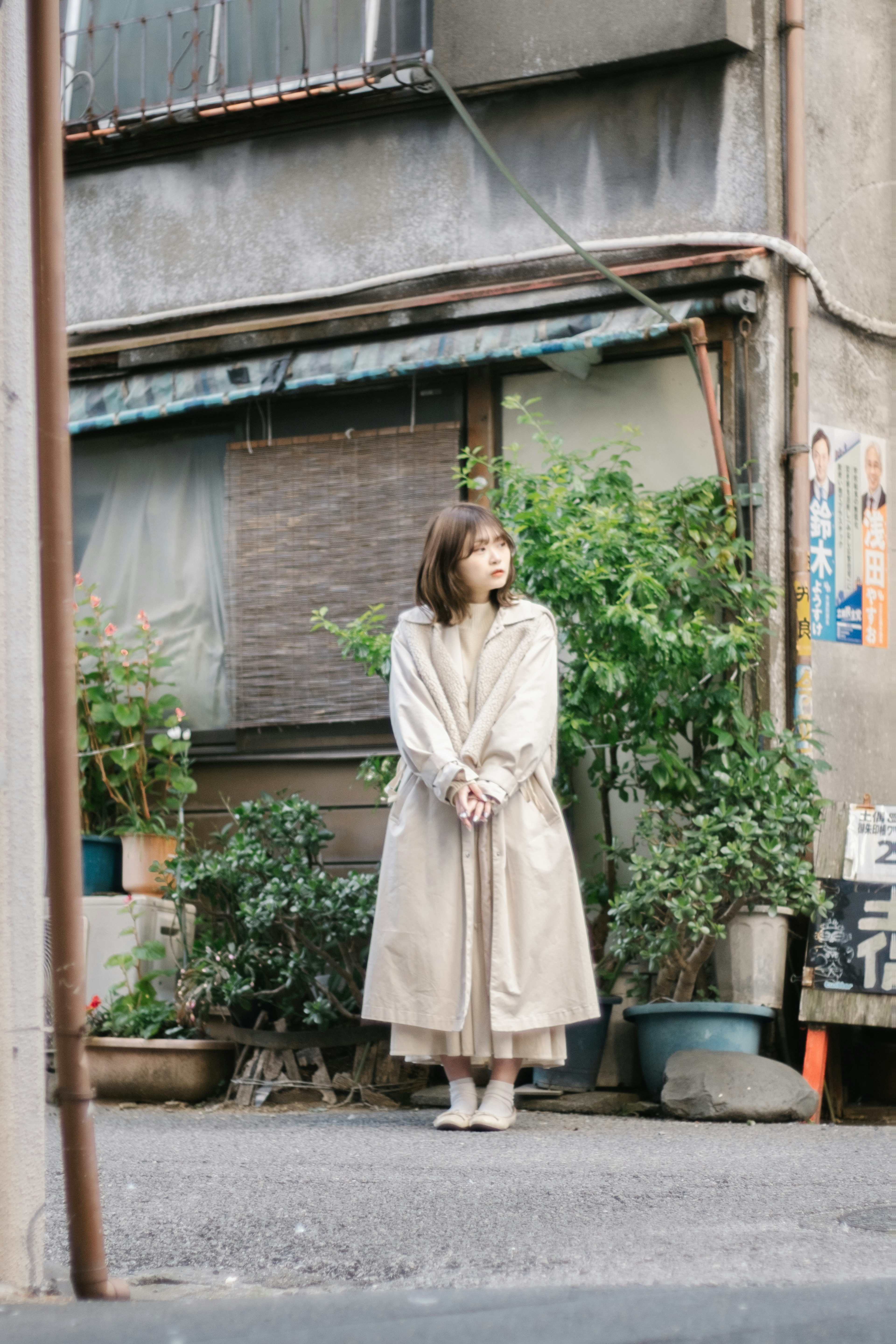 A woman standing at a street corner wearing a beige coat surrounded by green plants