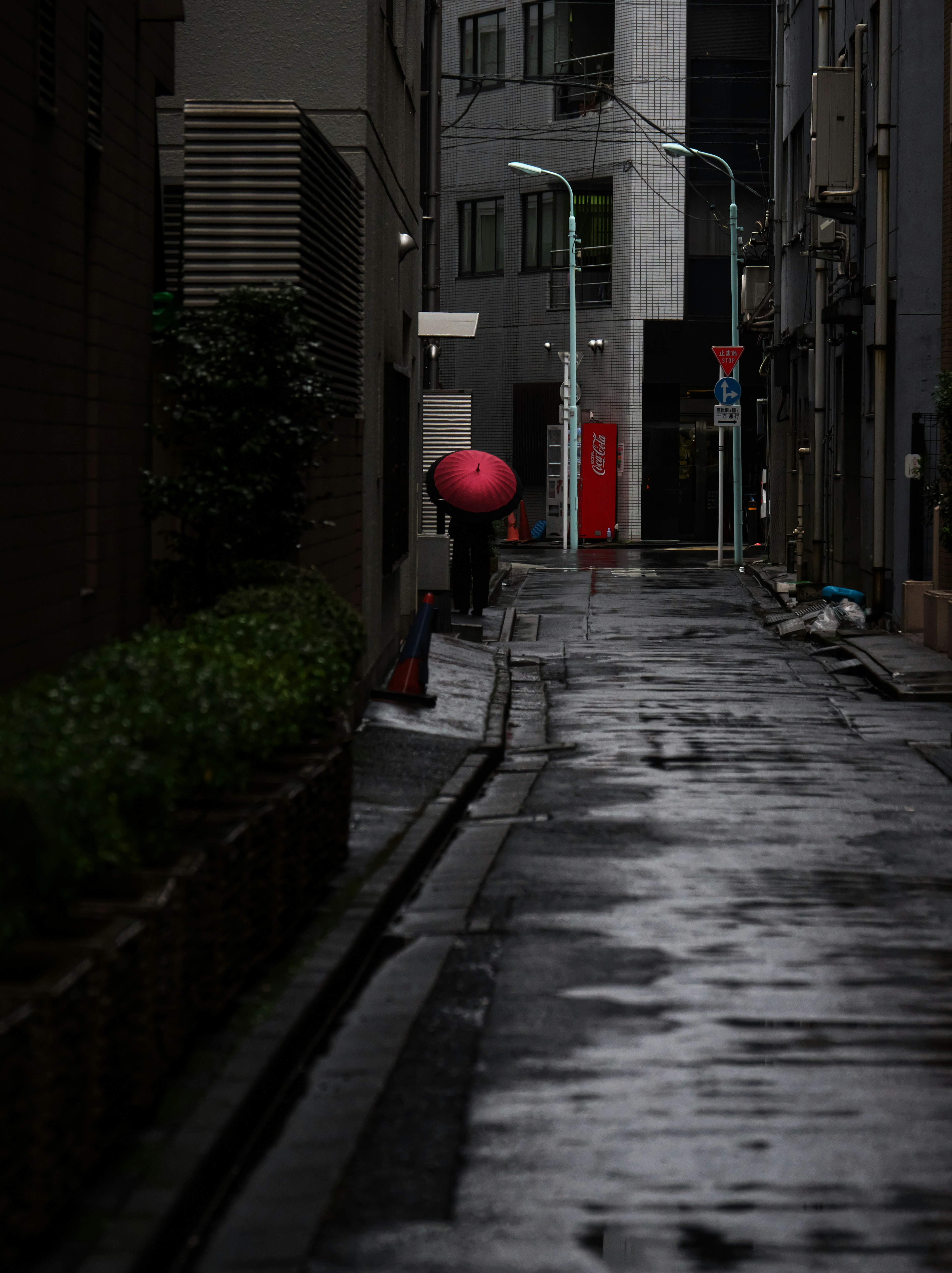 雨に濡れた狭い路地に赤い傘を持った人物が立っている