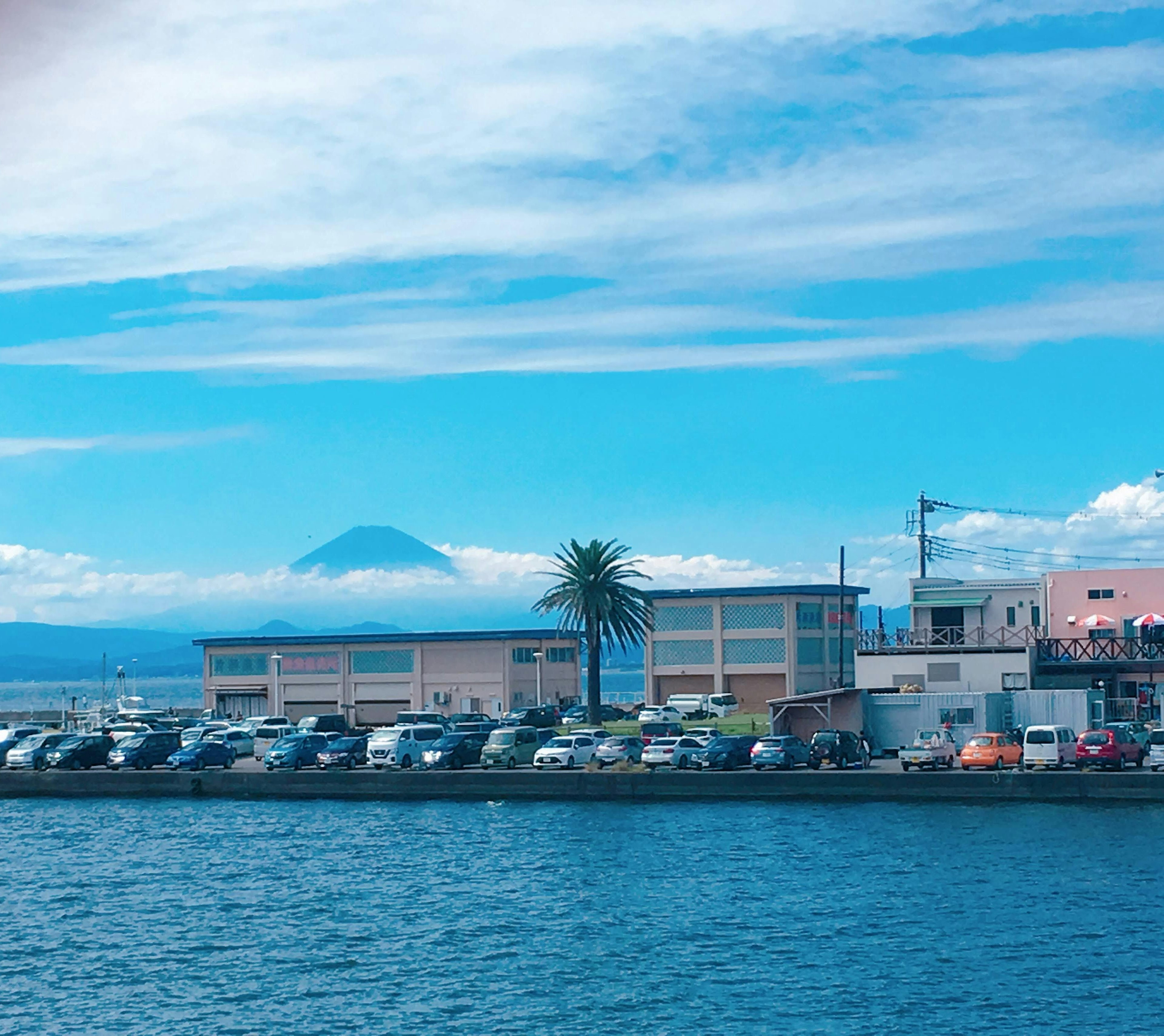 Pemandangan tepi laut dengan bangunan dan mobil terparkir di latar belakang langit biru dan Gunung Fuji