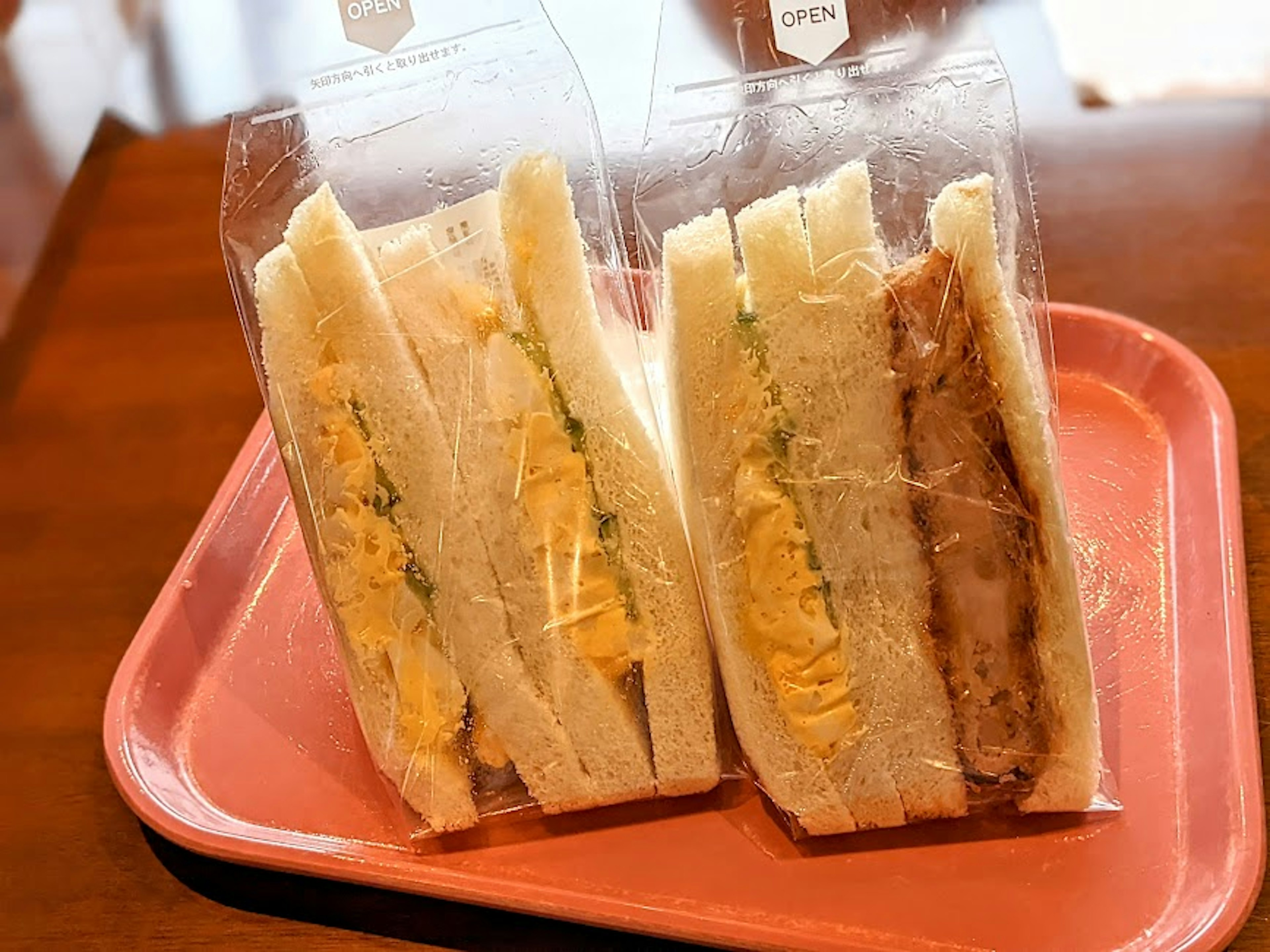 Dua sandwich dalam kemasan transparan di atas nampan merah muda