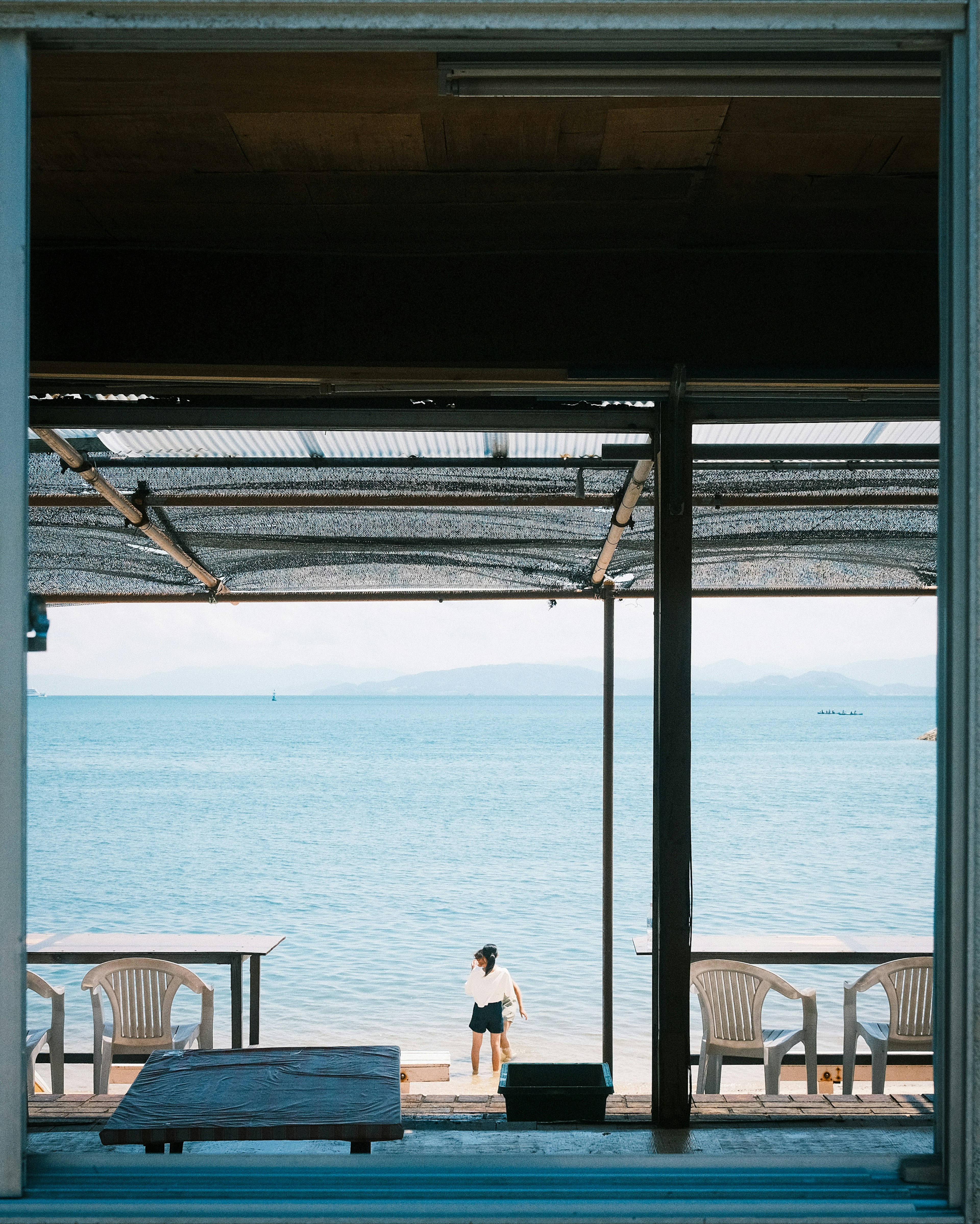 Persona de pie junto al mar vista a través de una ventana con sillas