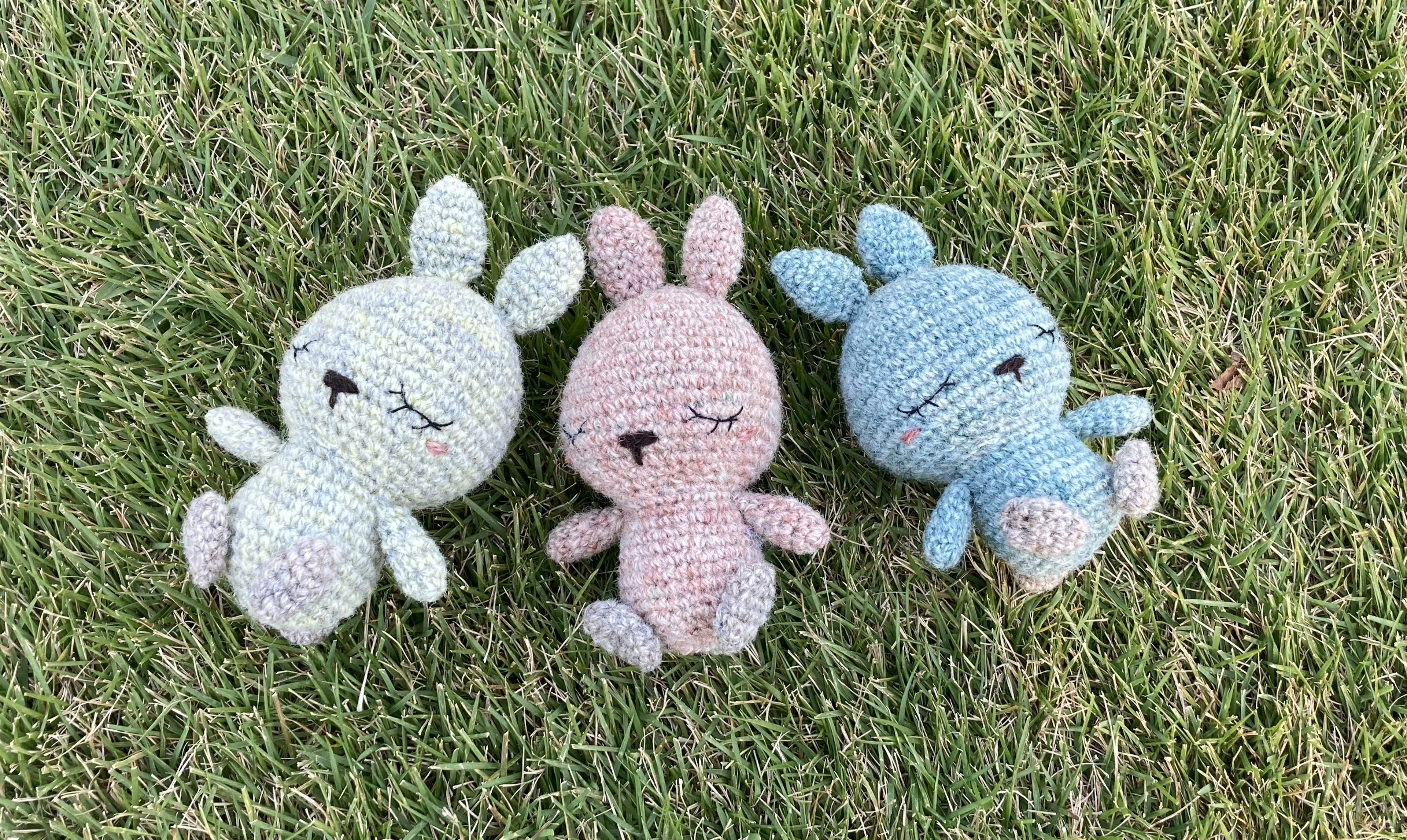 Three crochet bunny plush toys in green, pink, and blue lying on grass