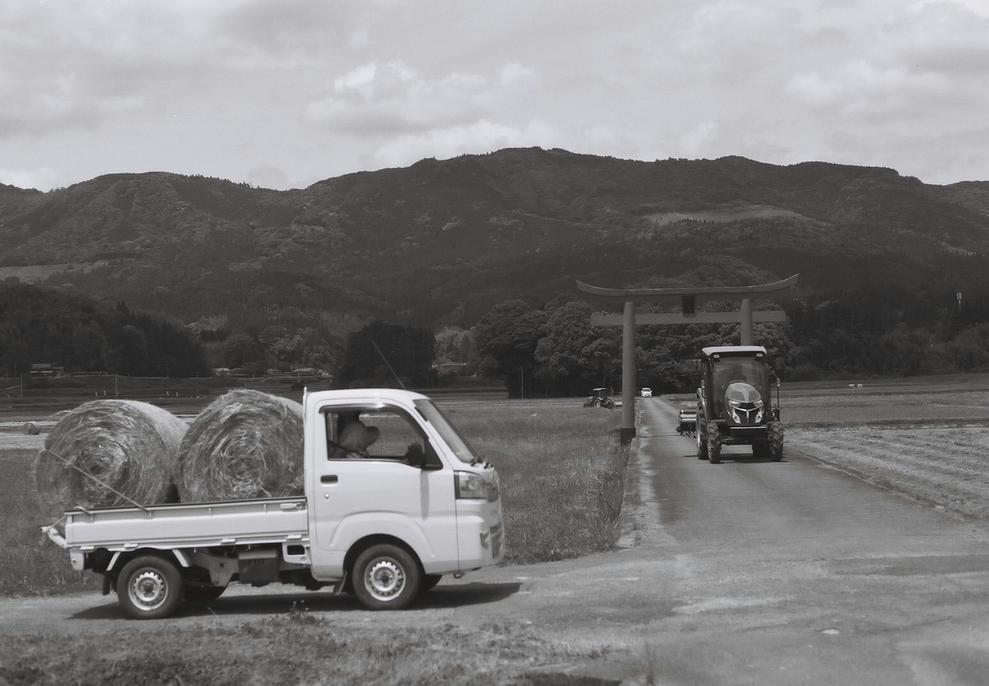 Escena rural en blanco y negro con una pequeña camioneta transportando fardos de heno por un camino rural