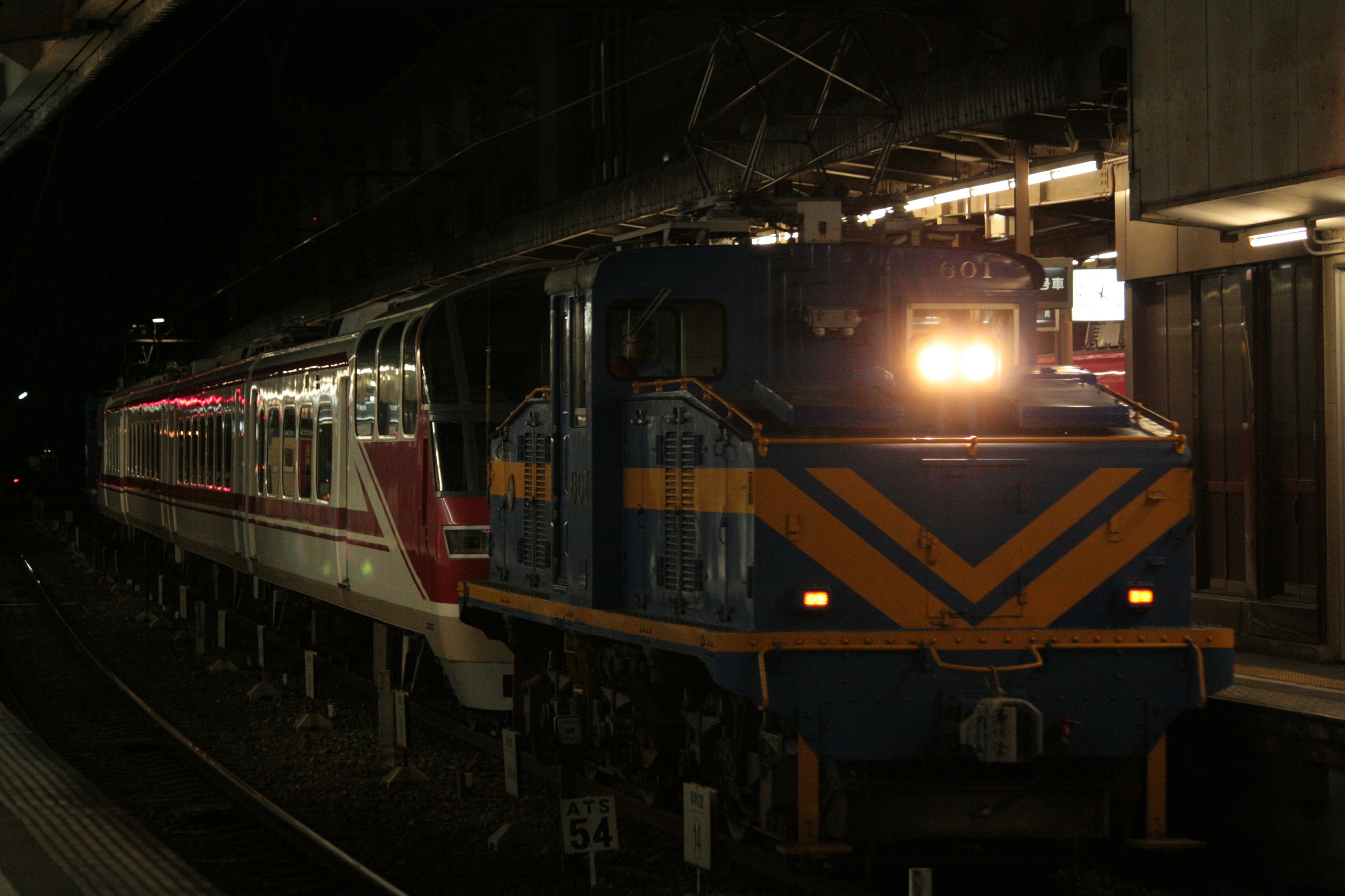 A blue and orange locomotive at a station with a red train during nighttime