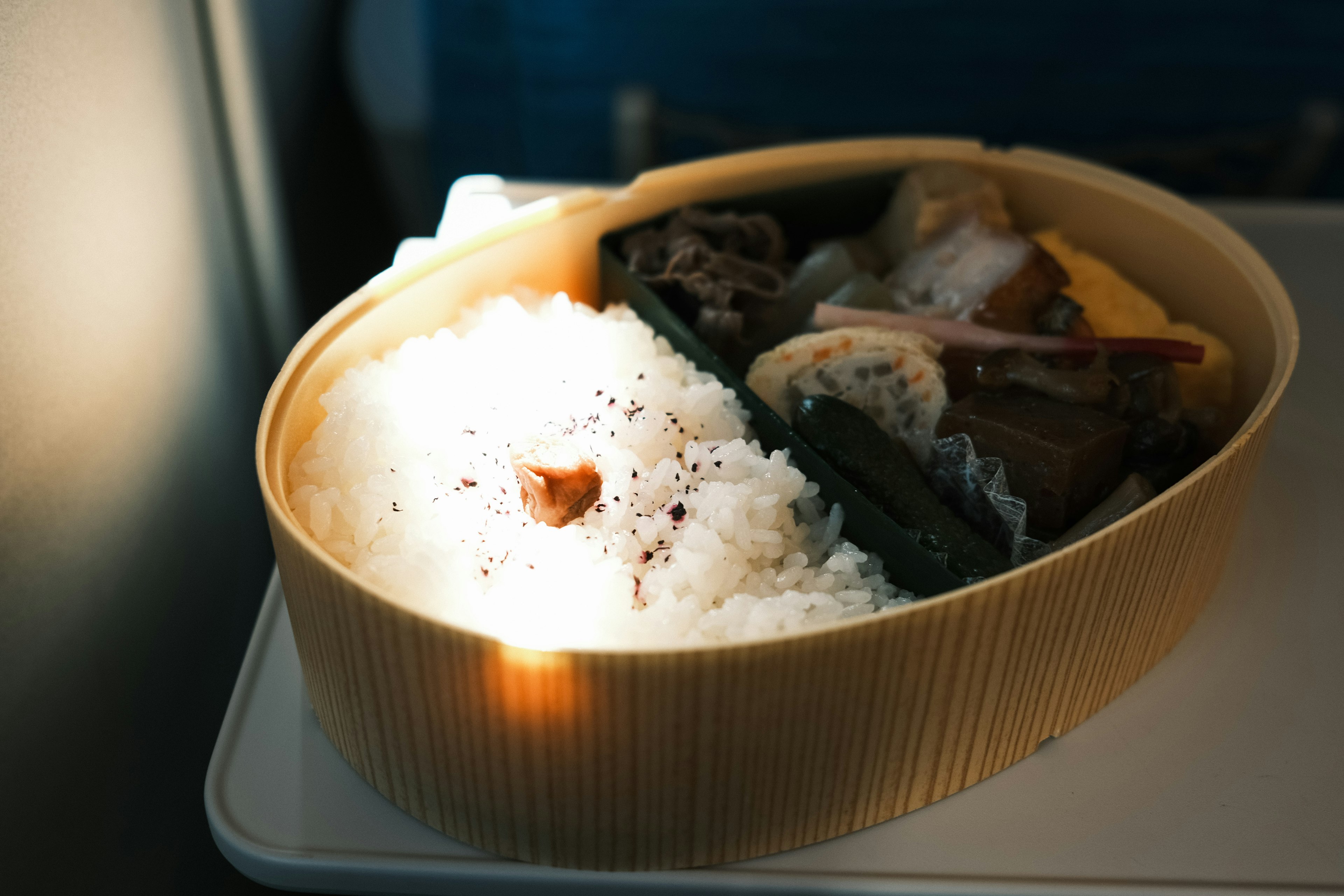 A photo of a bento box taken inside an airplane featuring rice and various side dishes in a bamboo container