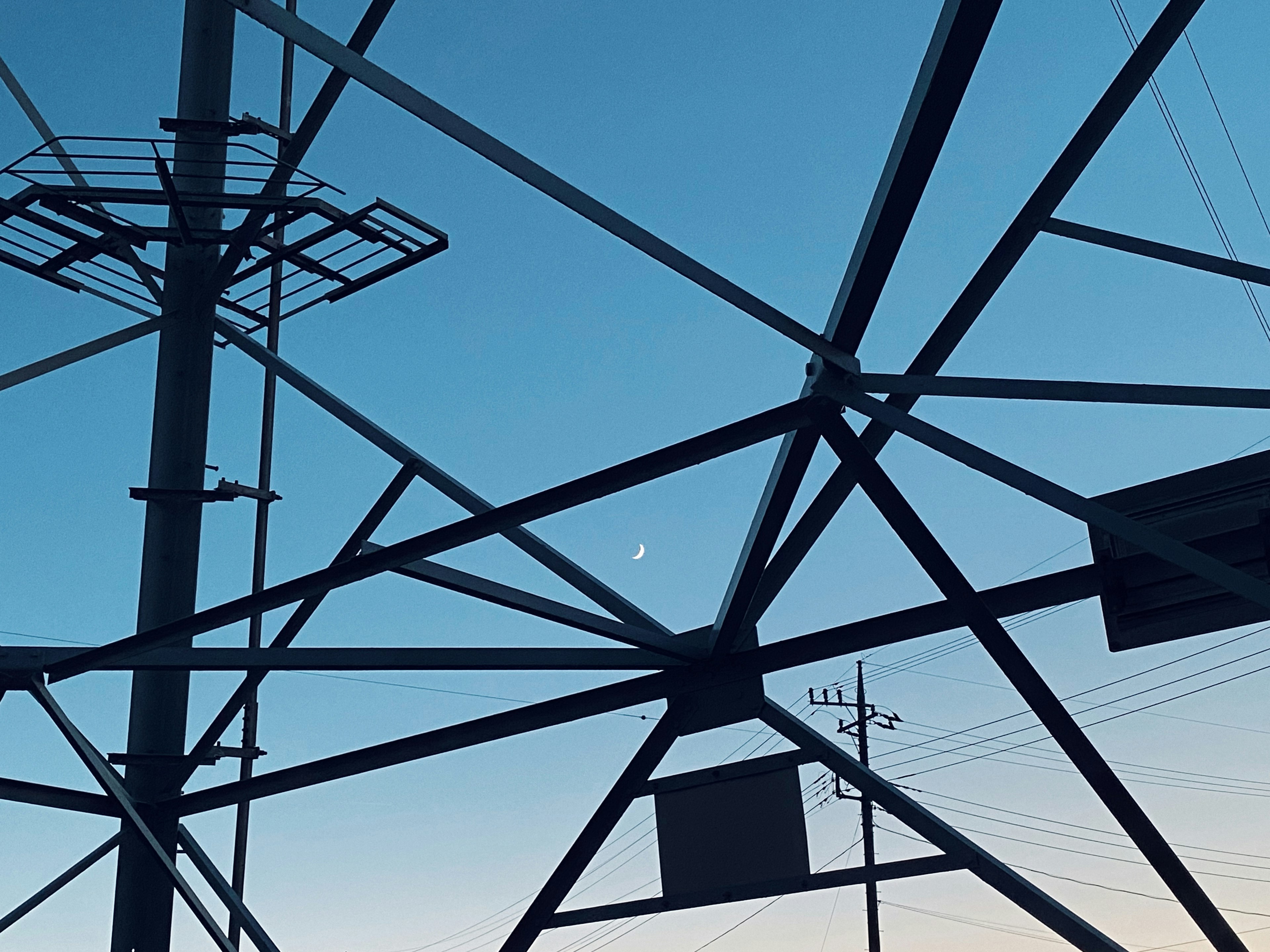 Une lune croissante visible parmi des lignes électriques et des structures en acier sur un ciel bleu