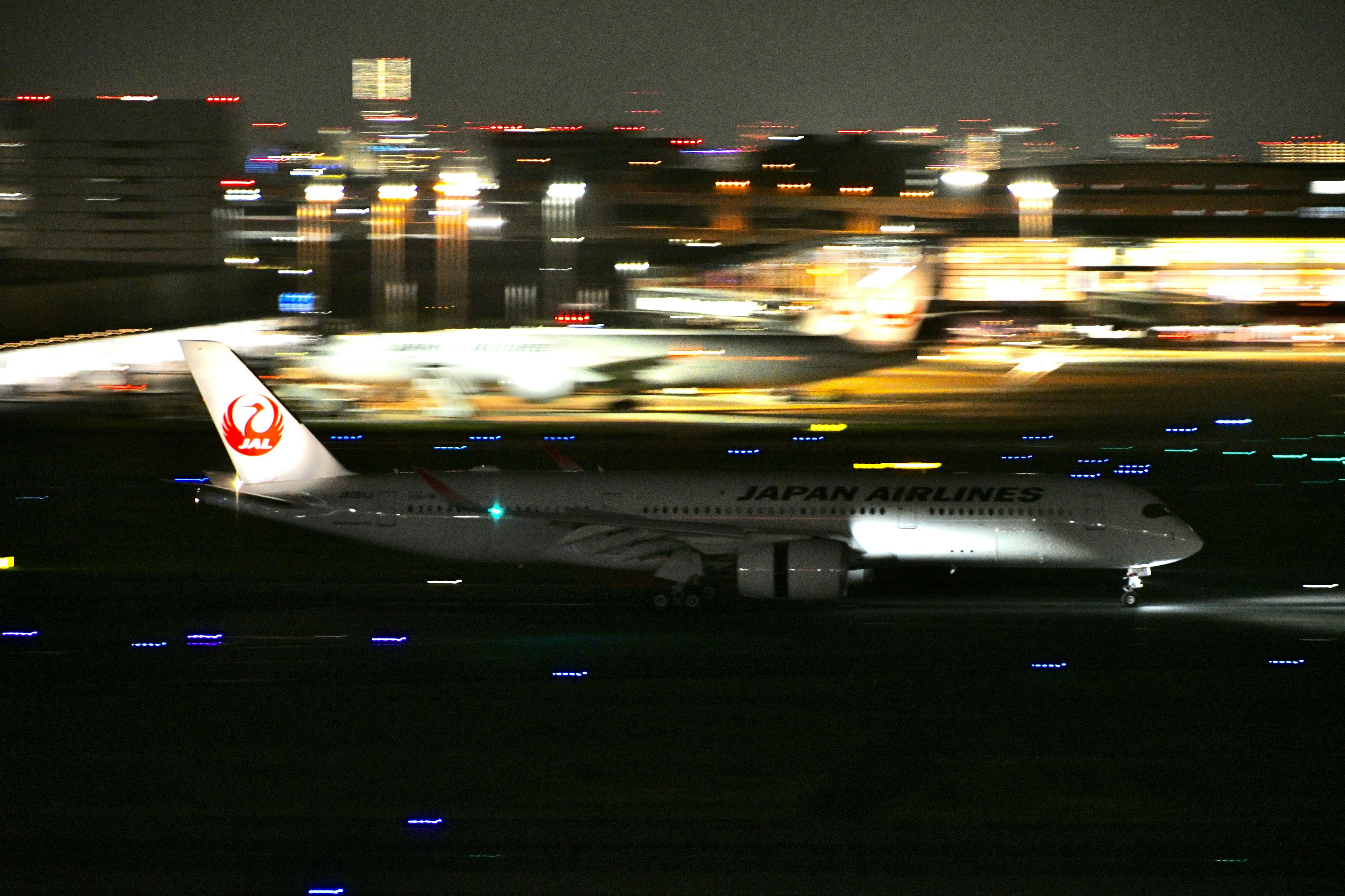 Pesawat Japan Airlines lepas landas di malam hari dengan lampu kota yang buram di latar belakang