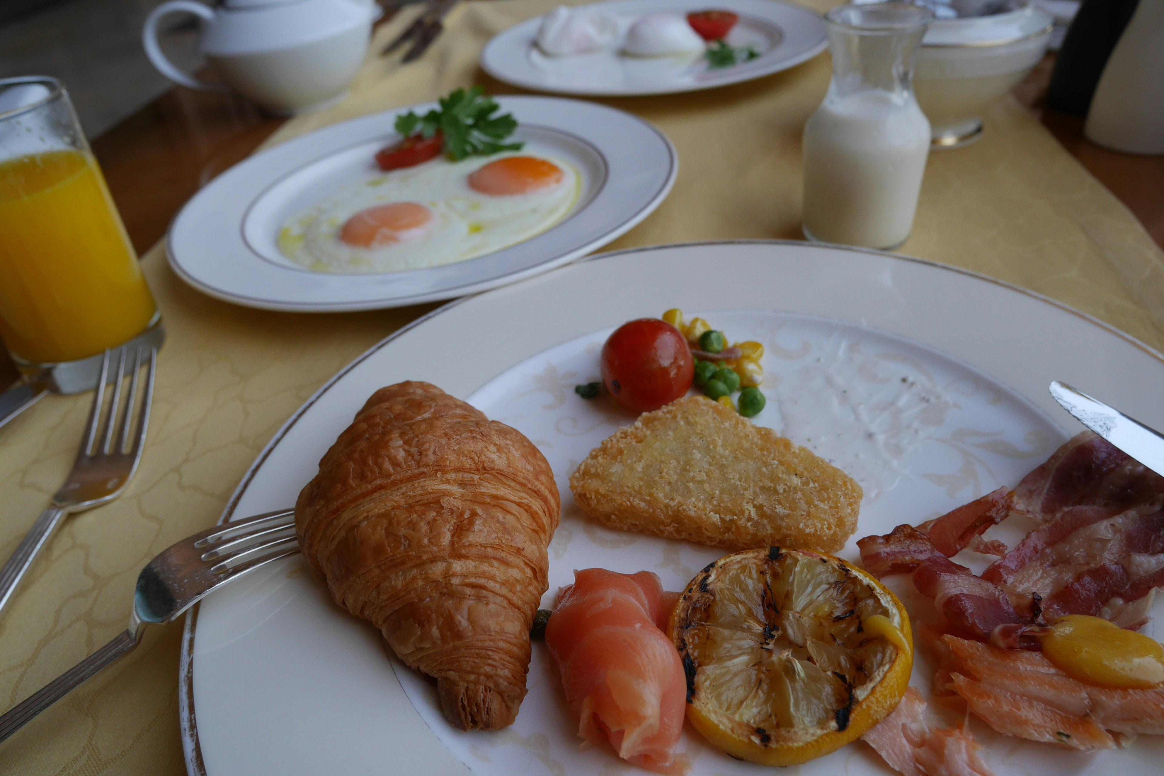 Piring sarapan dengan croissant ham tomat dan piring telur di latar belakang