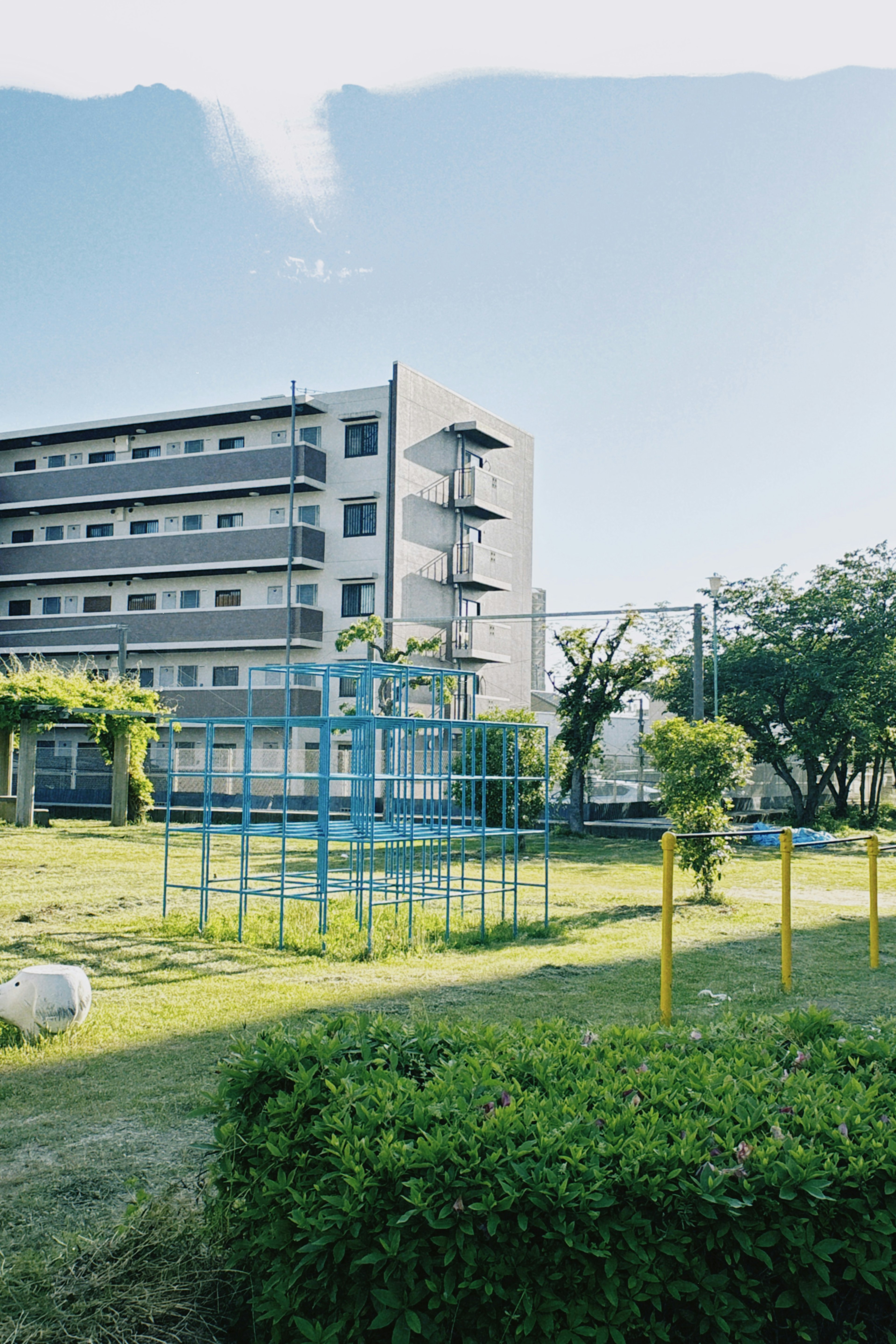 Pemandangan taman dengan peralatan bermain biru dan bangunan di dekatnya