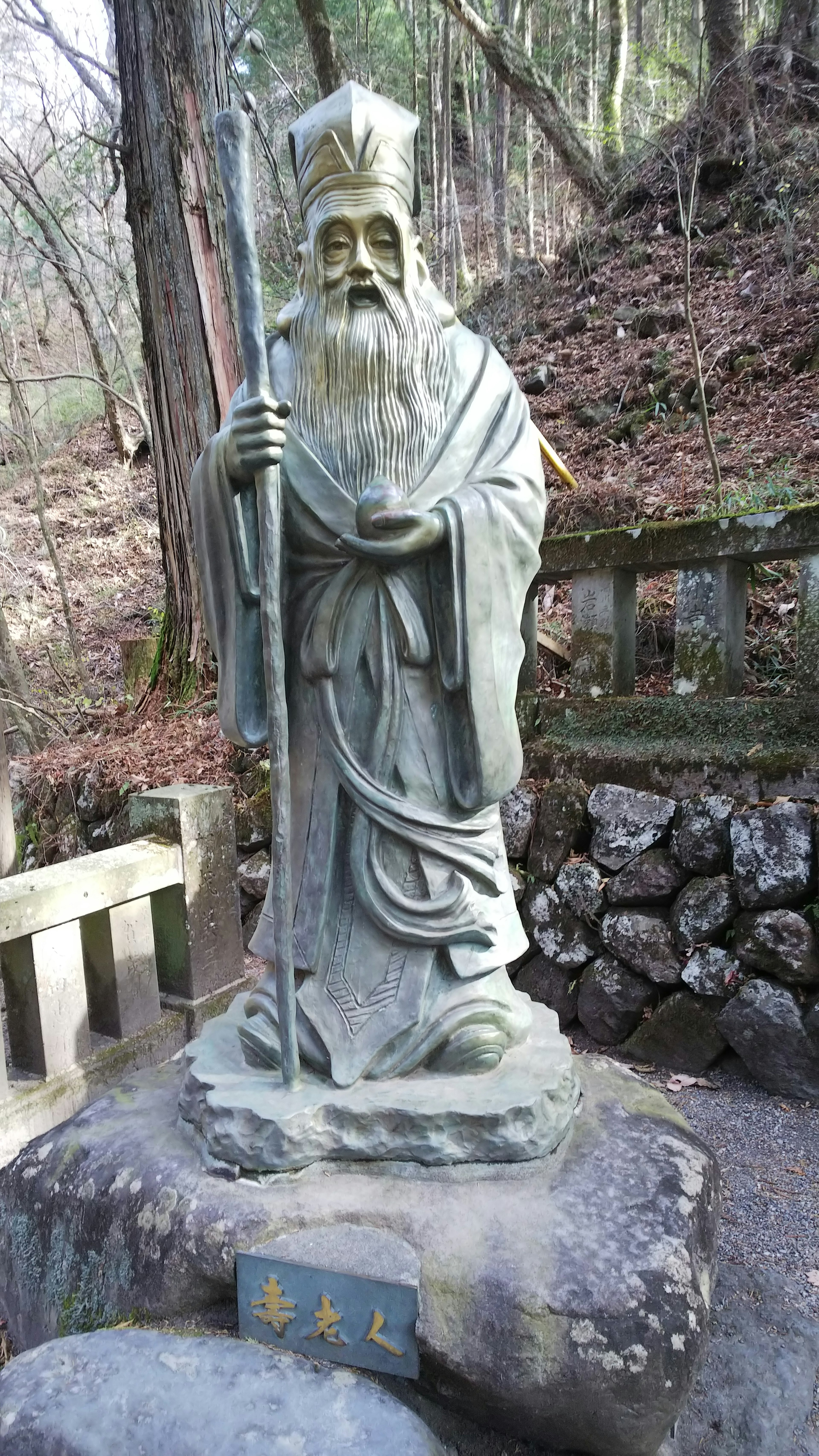 Una statua di pietra antica di una figura anziana con un bastone in un ambiente forestale