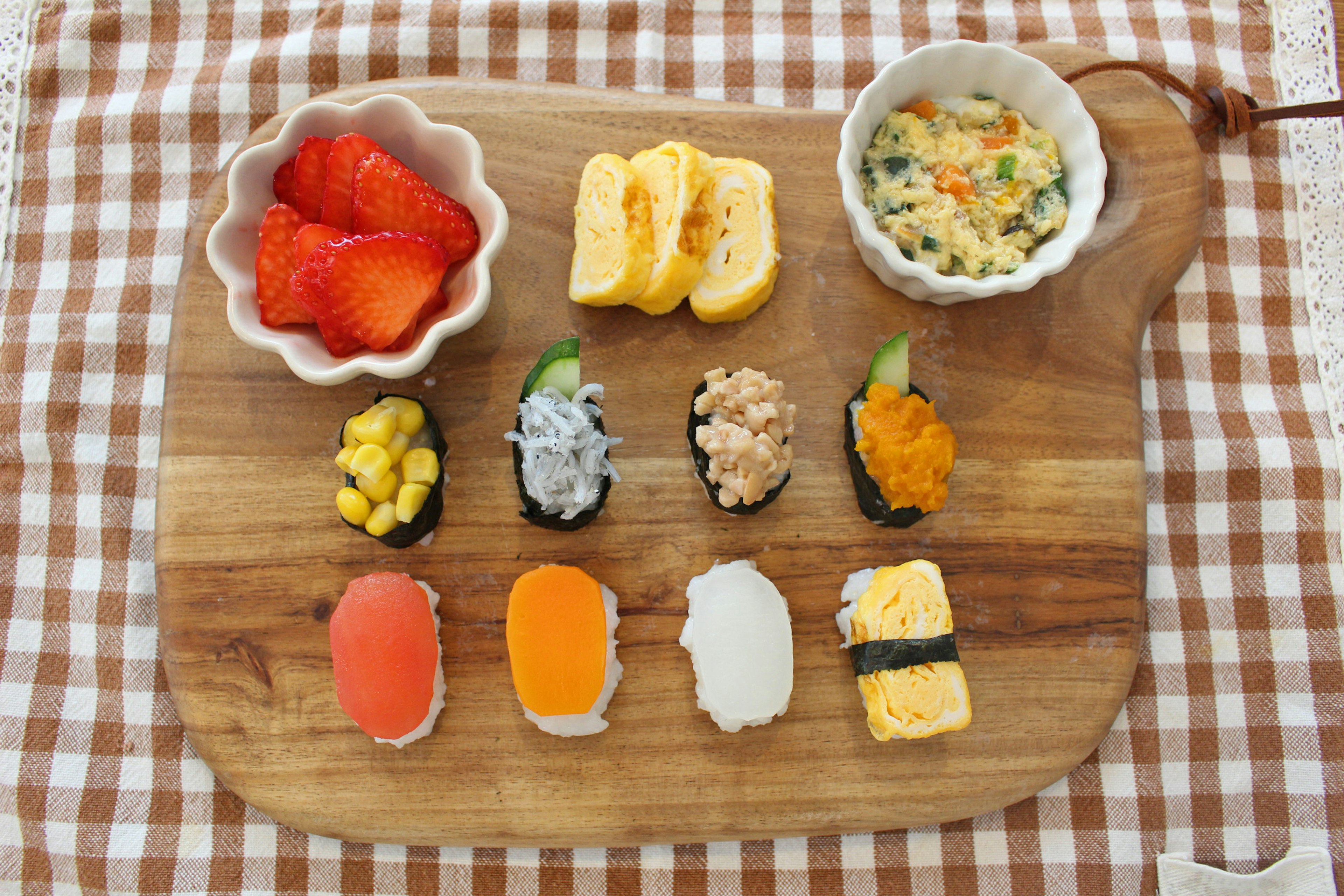 A wooden board displaying various sushi pieces and a bowl of strawberries