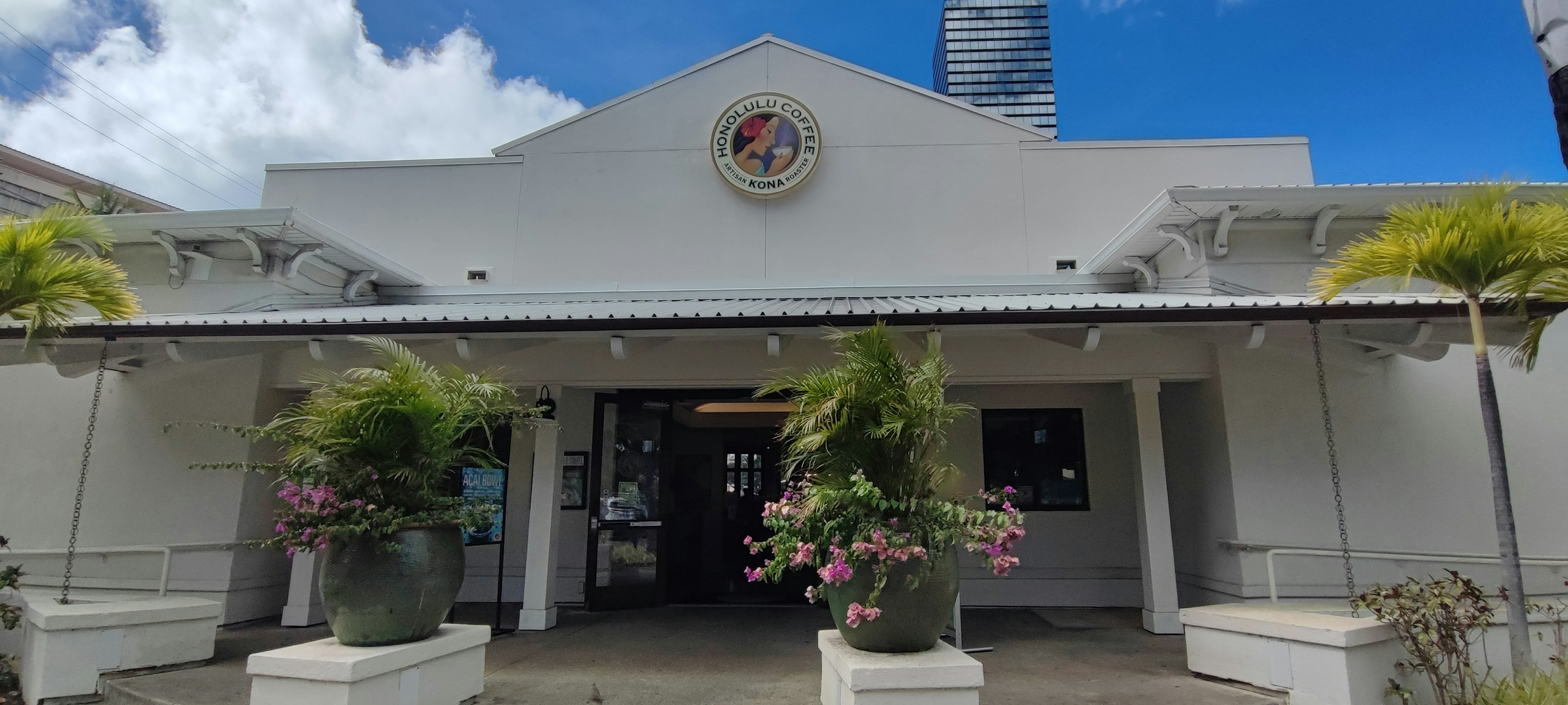 Fachada de un edificio blanco bajo un cielo azul con una entrada central y grandes macetas adornadas con flores