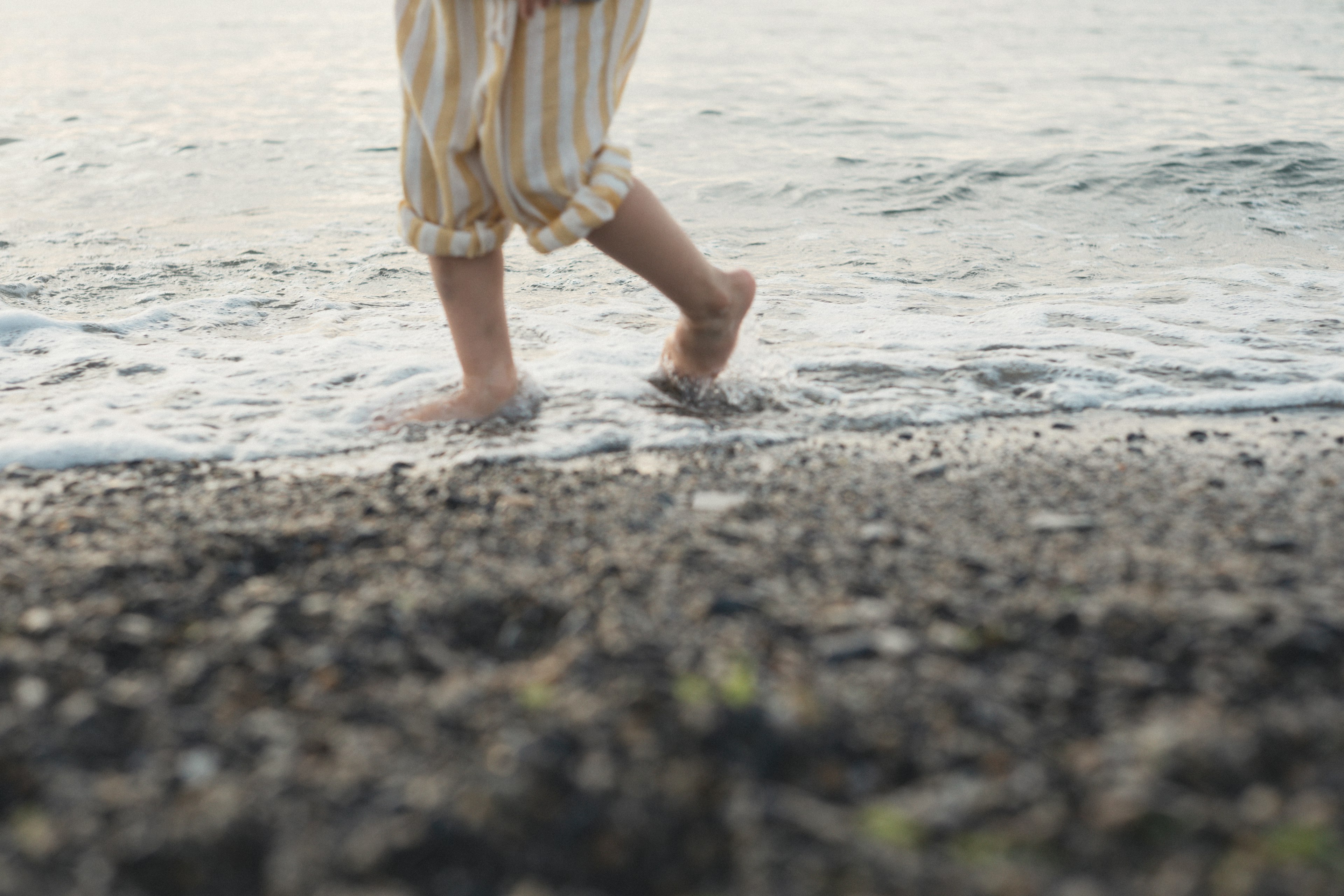 Füße eines Kindes in gestreiften Hosen, die im flachen Wasser am Strand laufen