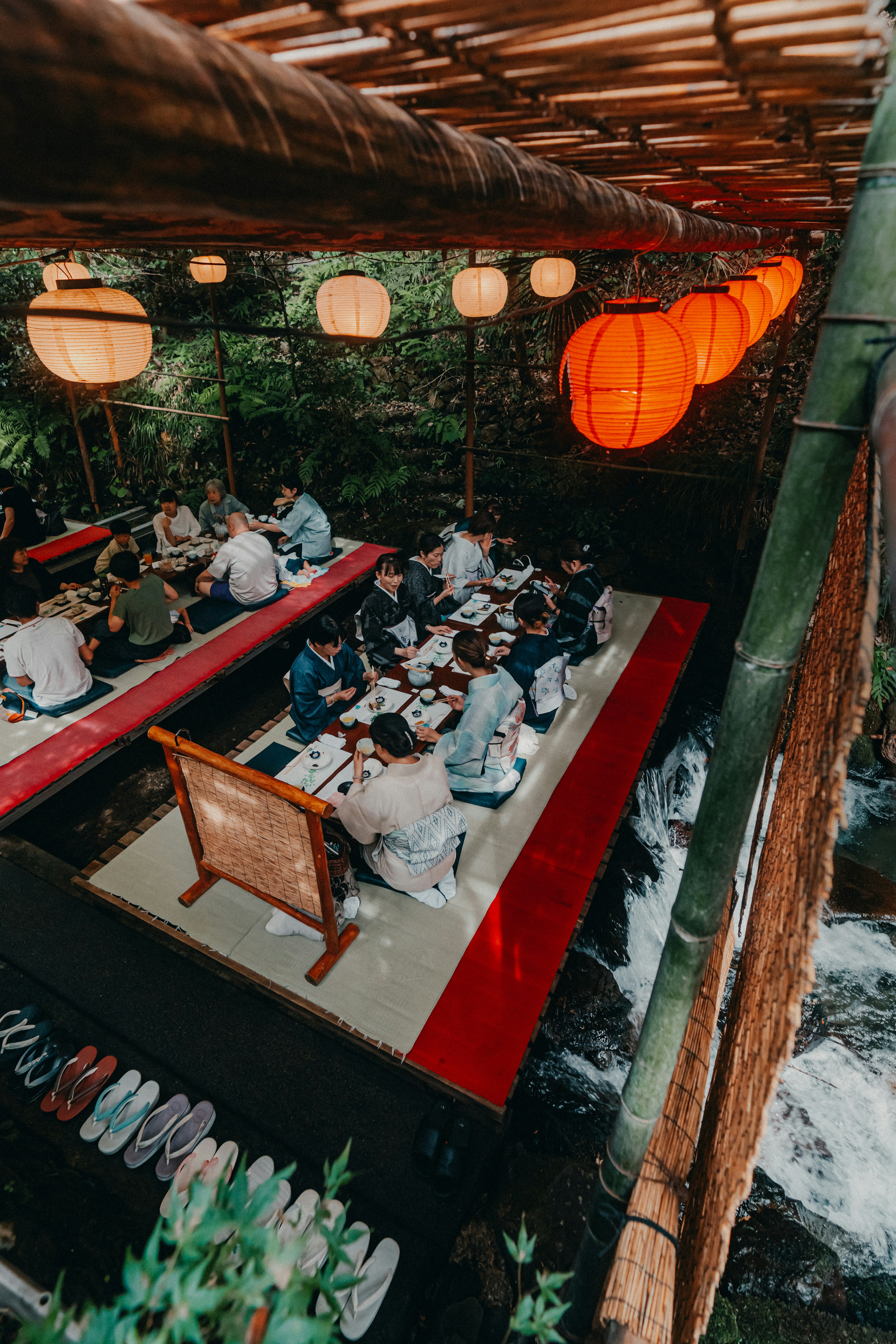 Personas cenando bajo un techo de bambú junto al río con faroles rojos