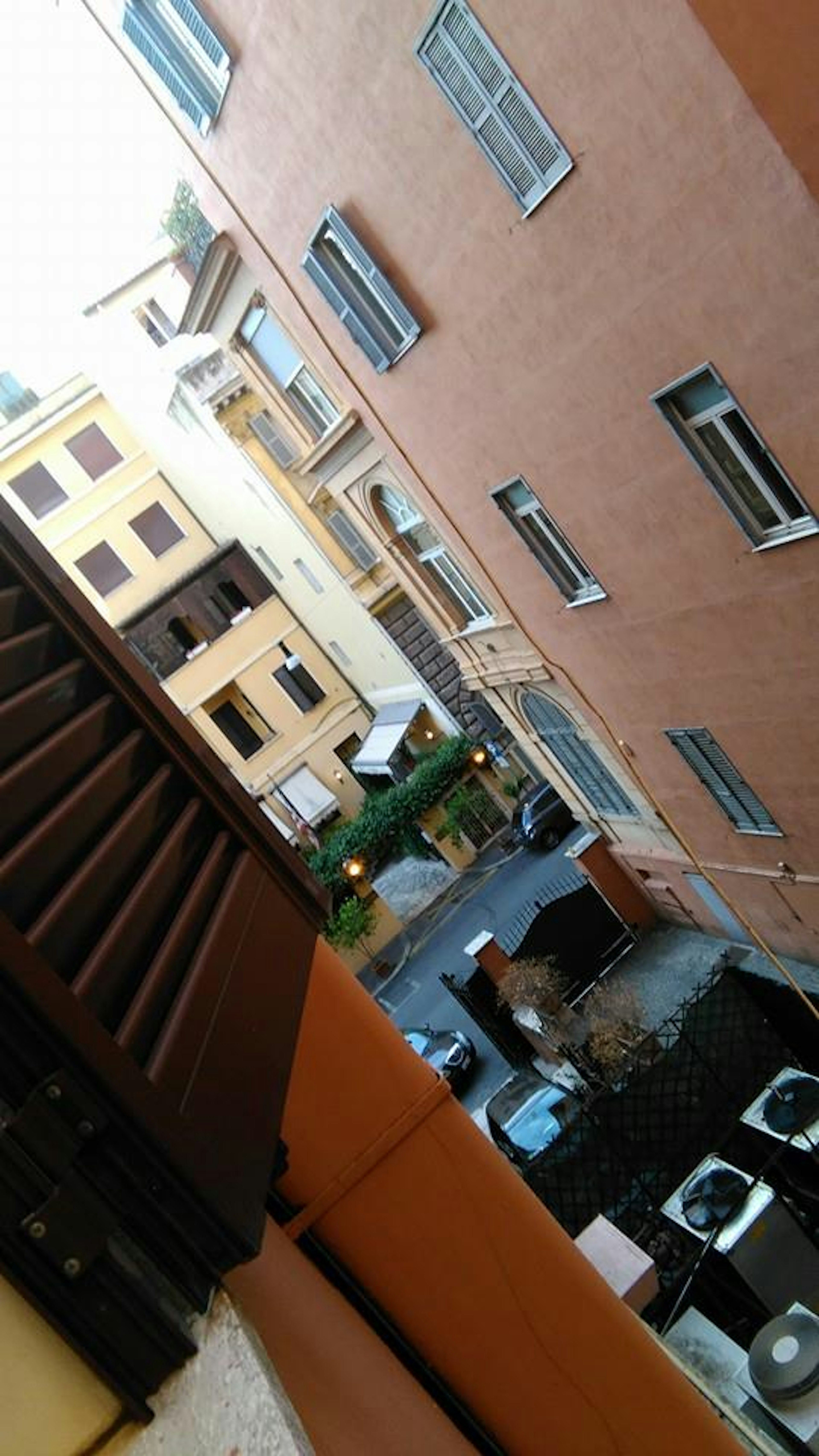 Vista da una finestra che mostra un paesaggio urbano con edifici in mattoni rossi strada stretta diverse auto e unità di condizionamento