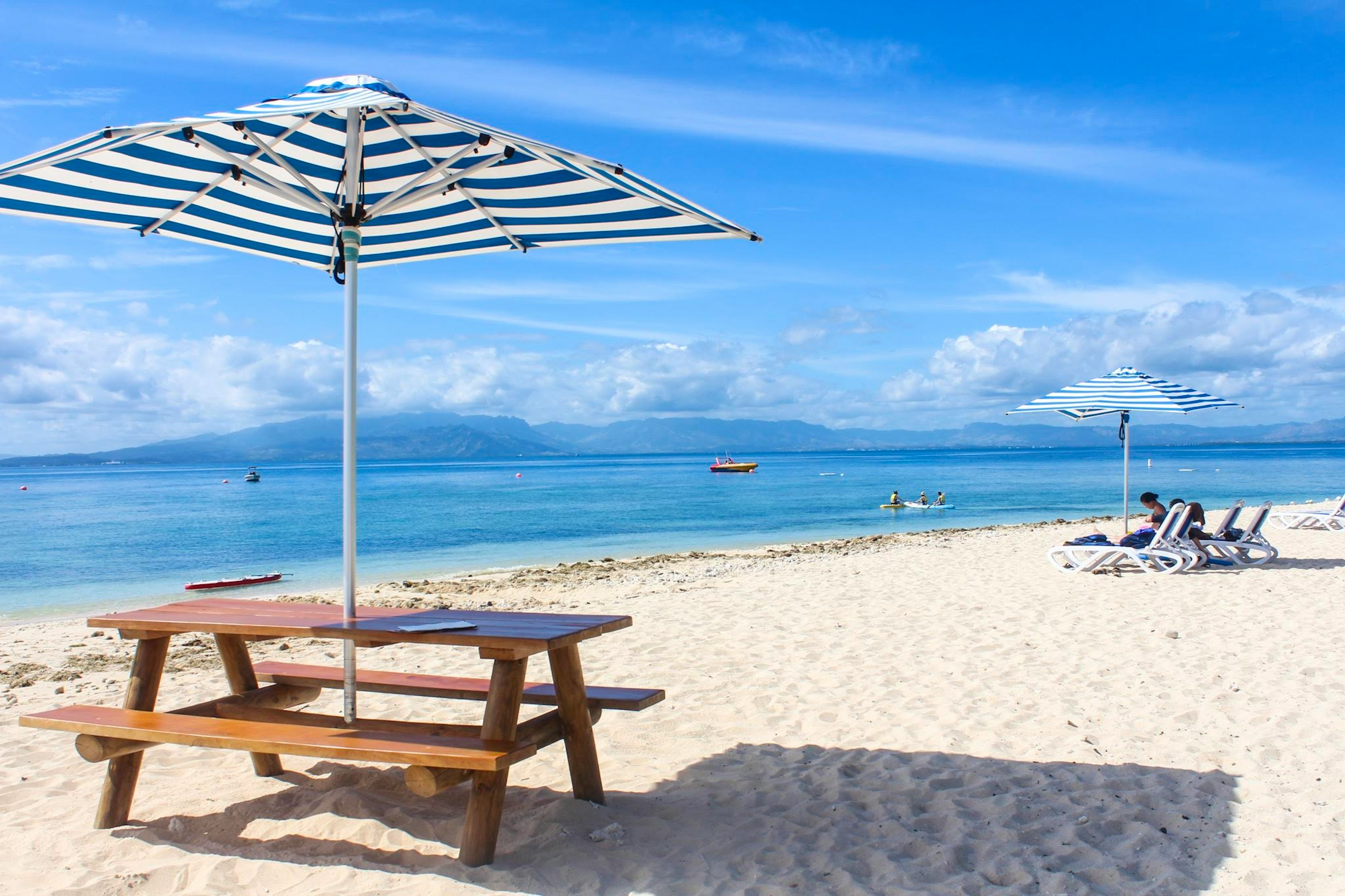 Holztisch mit gestreiftem Sonnenschirm an einem Strand mit weißem Sand und blauem Himmel