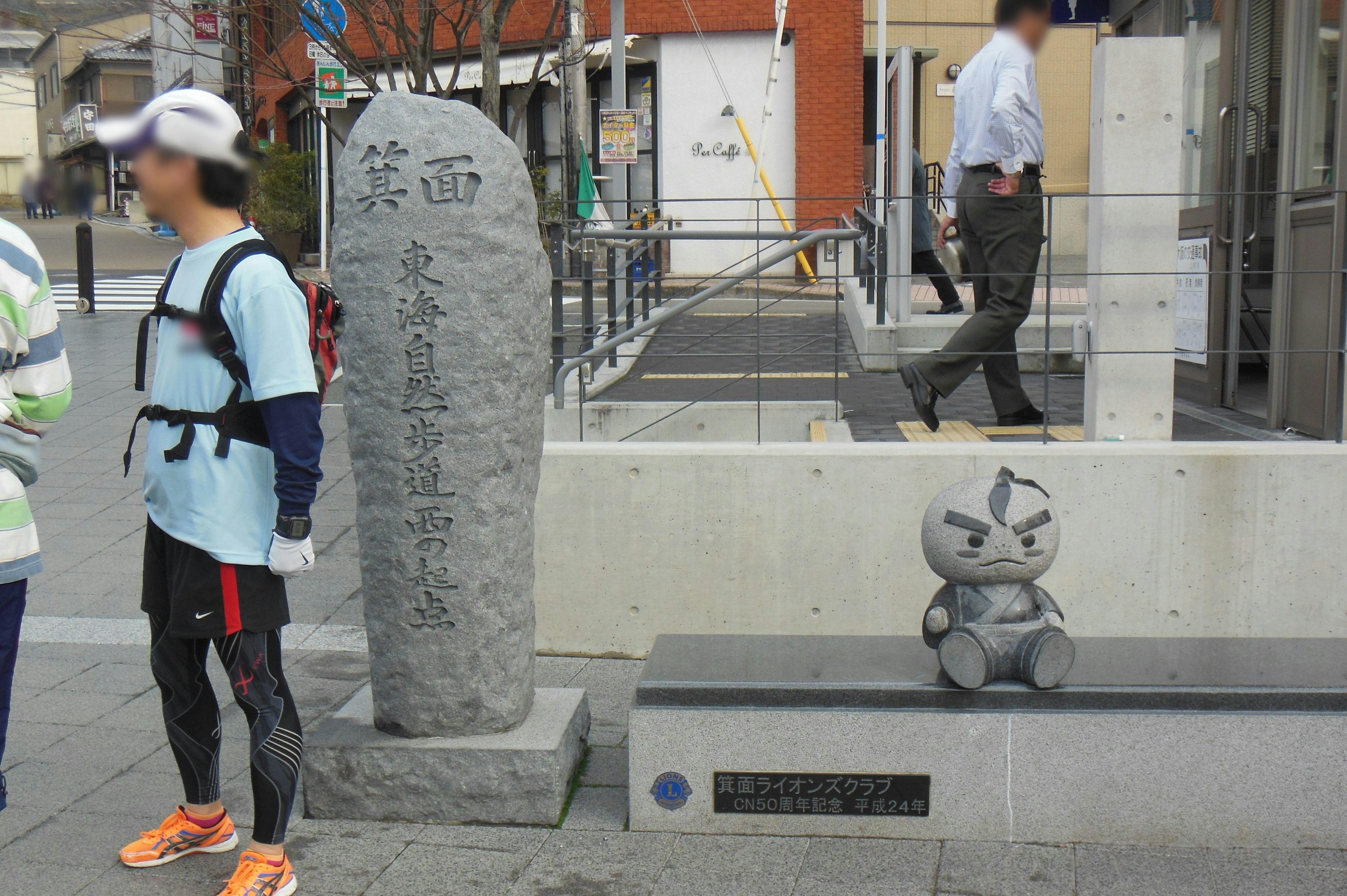 石碑と小さなキャラクターの像が並ぶ街角の風景