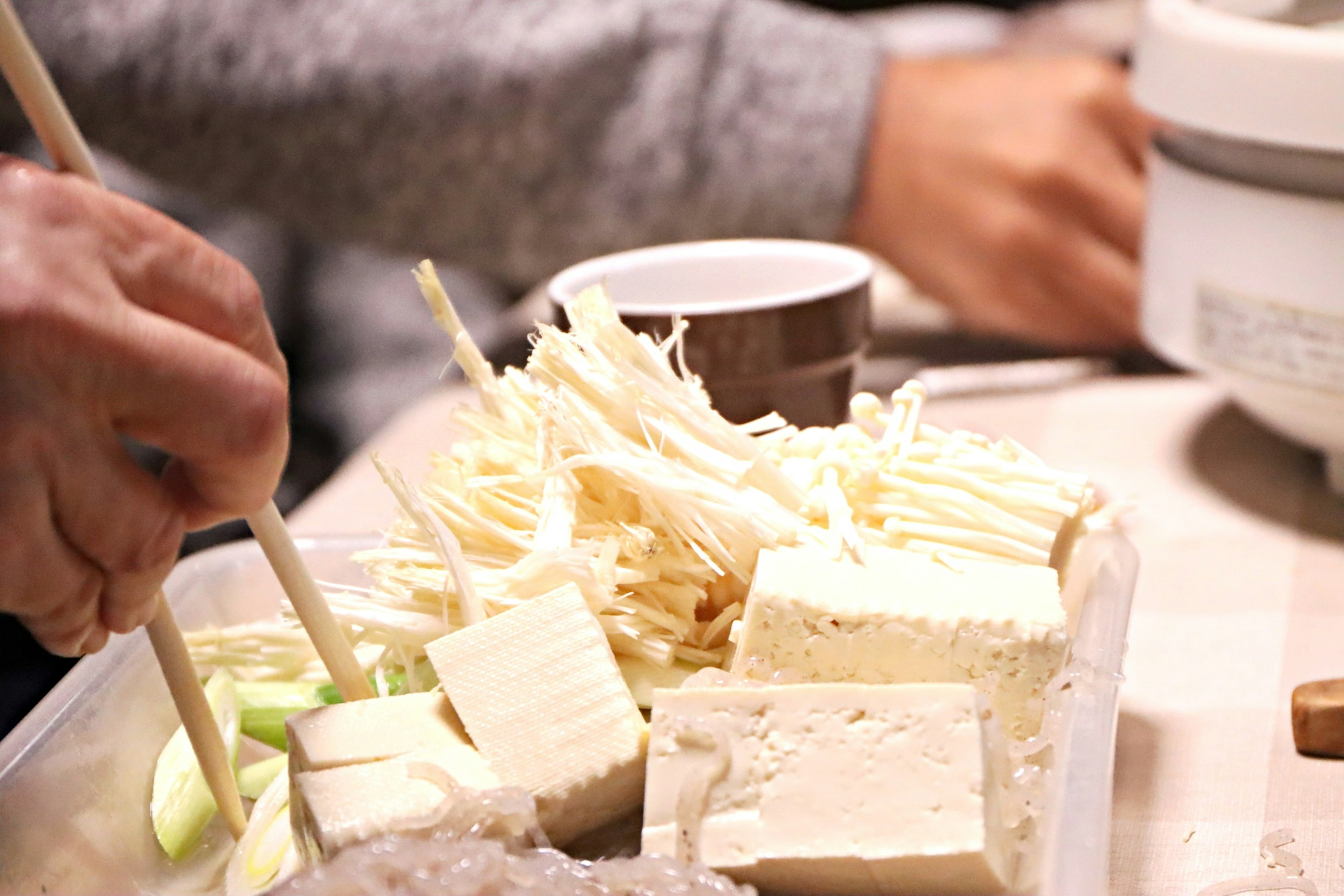 Hände, die Essstäbchen benutzen, um Tofu und Enoki-Pilze von einem Tablett zu nehmen