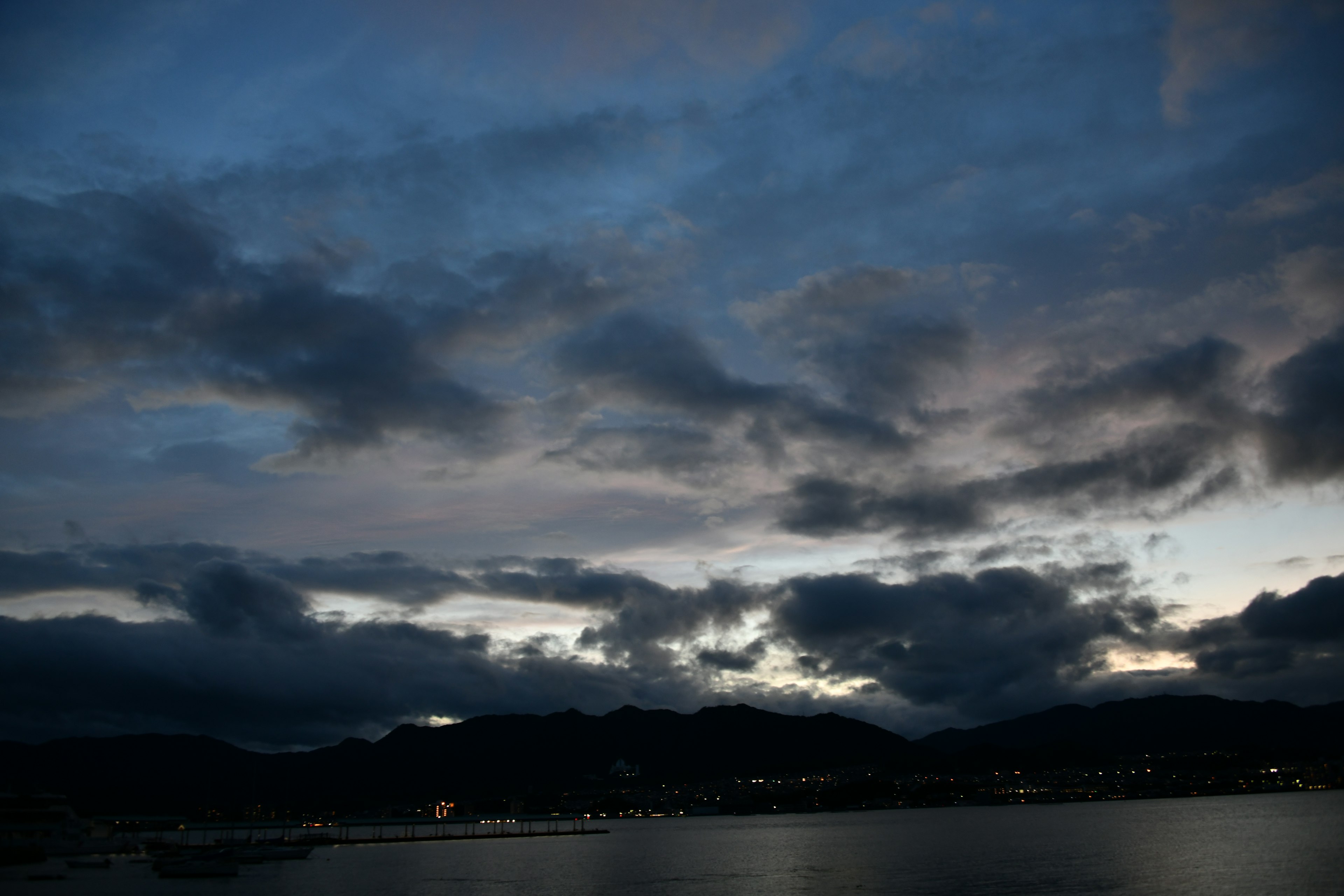 暗い空と雲が広がる風景に、遠くに明かりが見える海岸