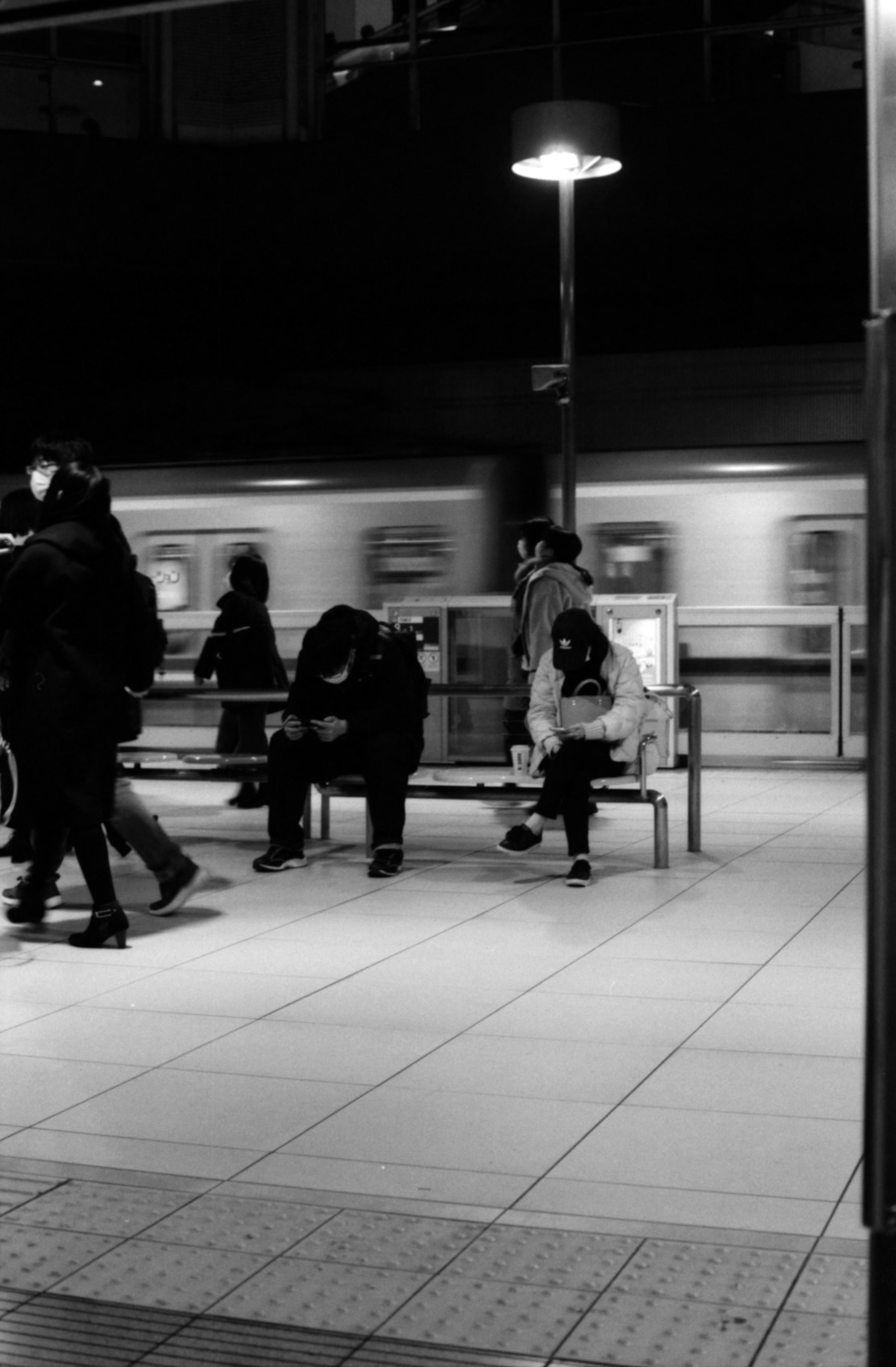 Photo en noir et blanc de personnes assises sur un banc à une station de train avec un train passant