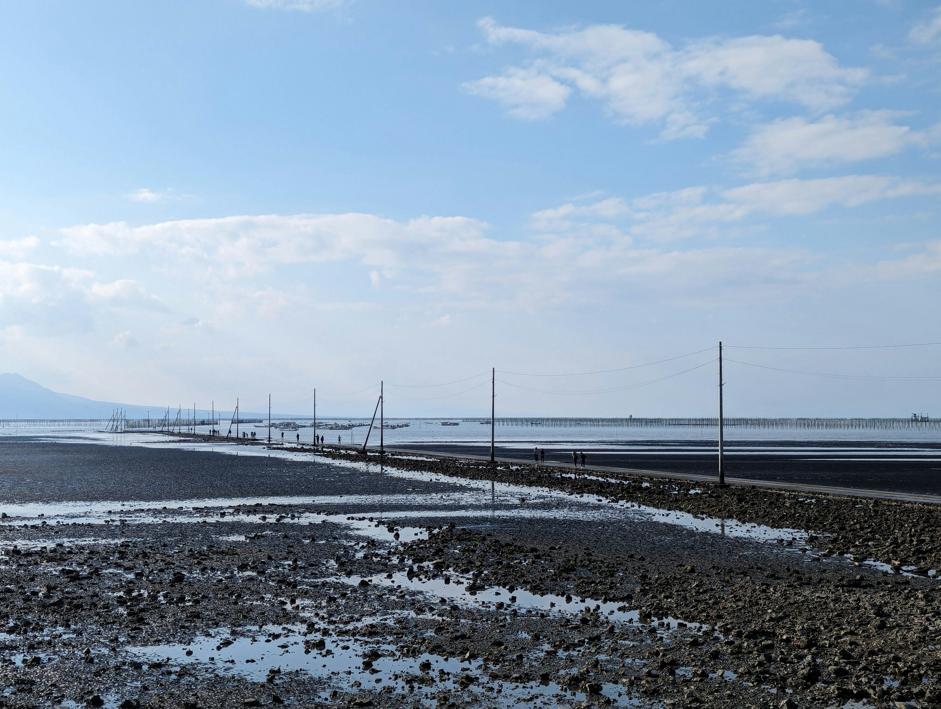 広い干潟と青い空が広がる風景で、干潟には数本のポールが立っている