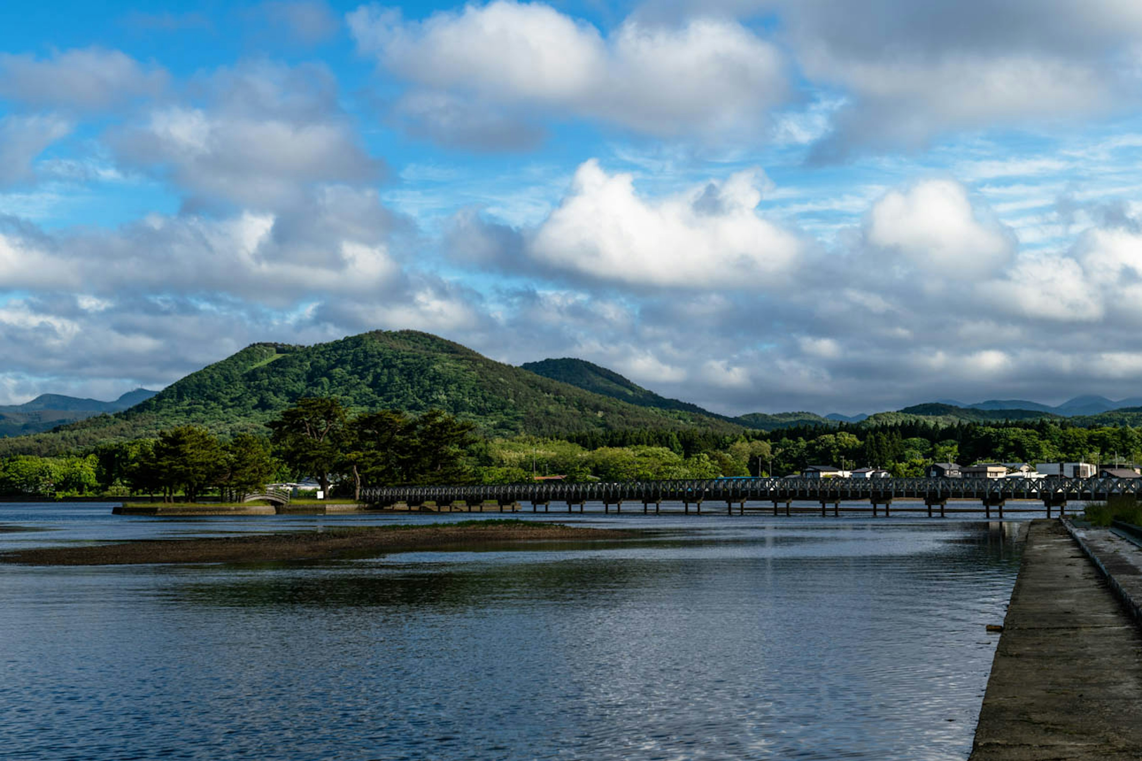 青い空と白い雲が浮かぶ風景 緑の山と静かな水面 橋と小さな町が見える