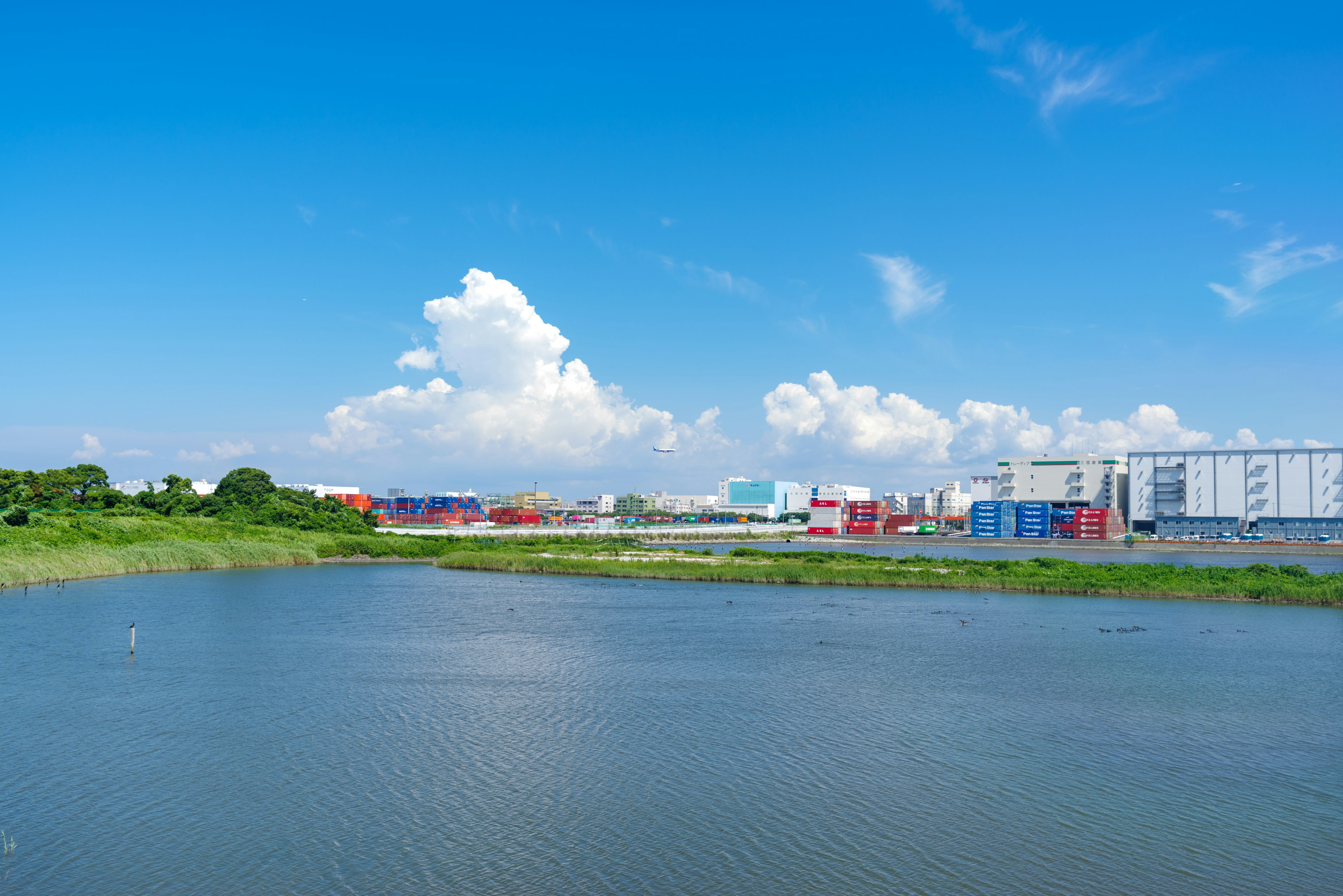 Landscape featuring a river under a blue sky with white clouds and industrial buildings