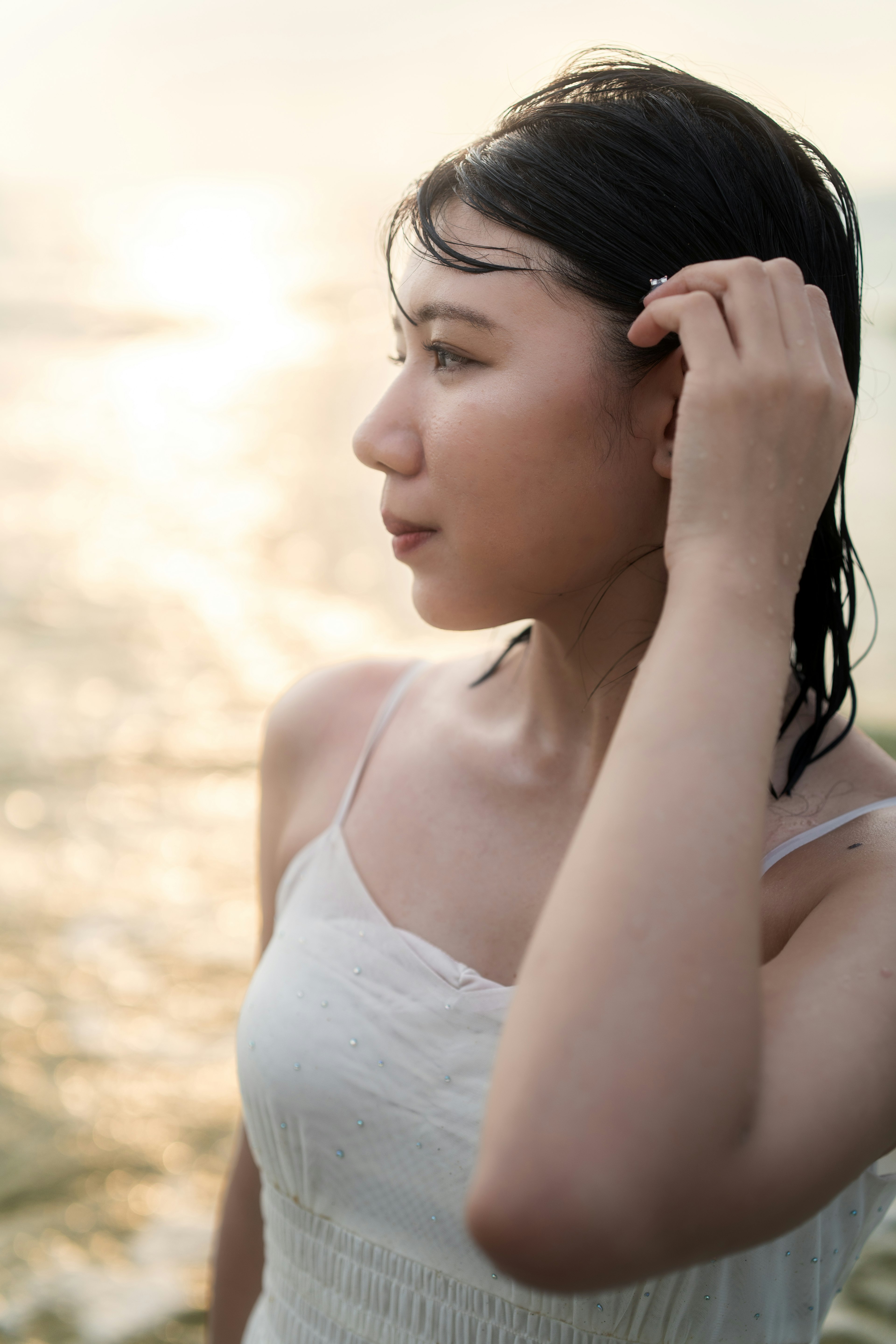 Side profile of a woman touching her hair by the beach wearing a light dress with shimmering water in the background