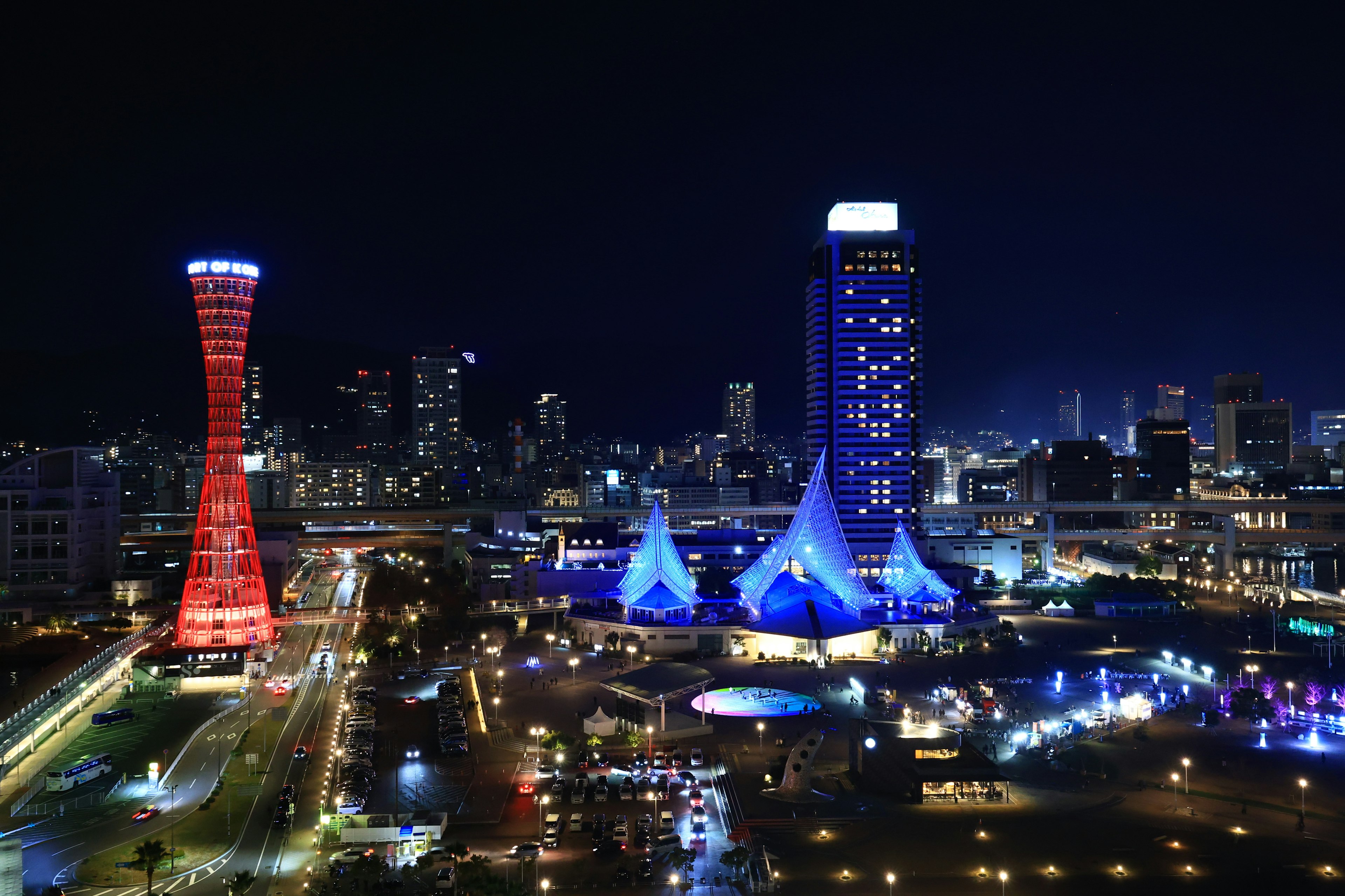 Vista nocturna de Kobe con una torre roja y carpas azules entre los rascacielos de la ciudad