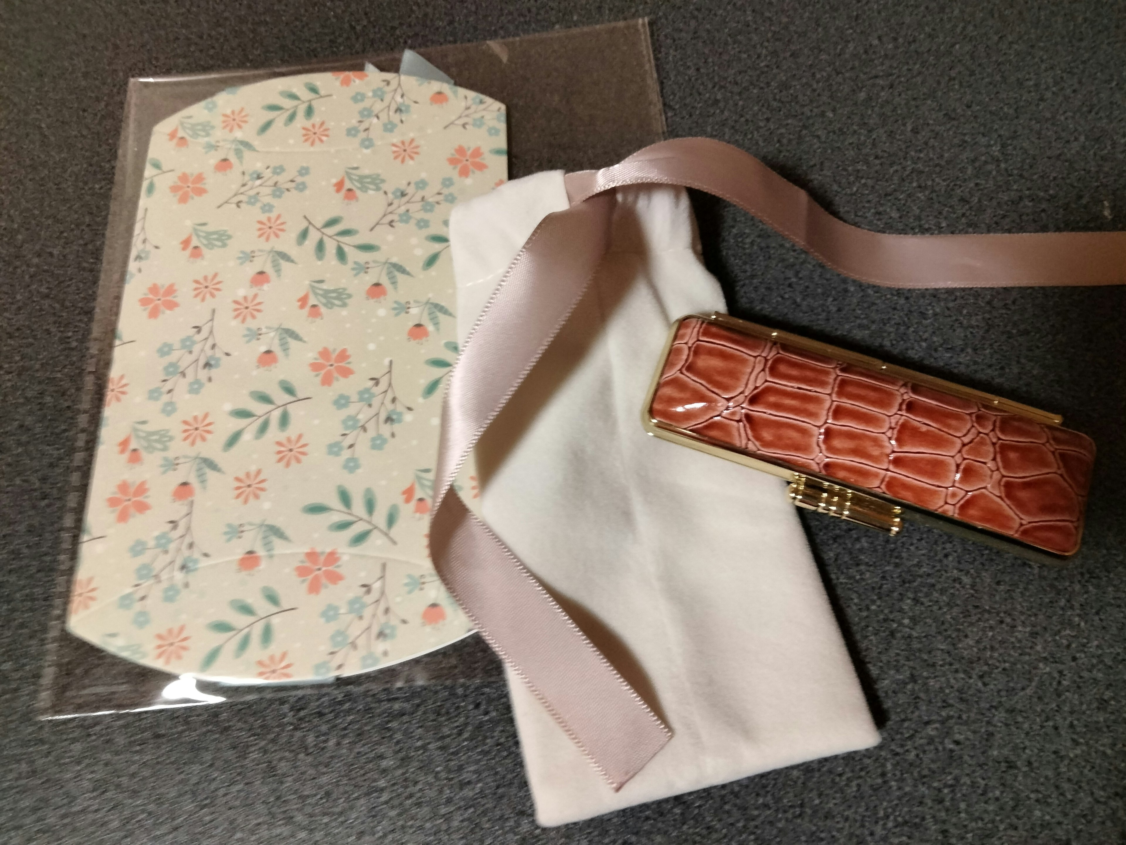 Ein florales Kuvert mit rosa Blumen und grünen Blättern neben einem weißen Stoffbeutel und einem goldenen Krokodil-Textur-Etui