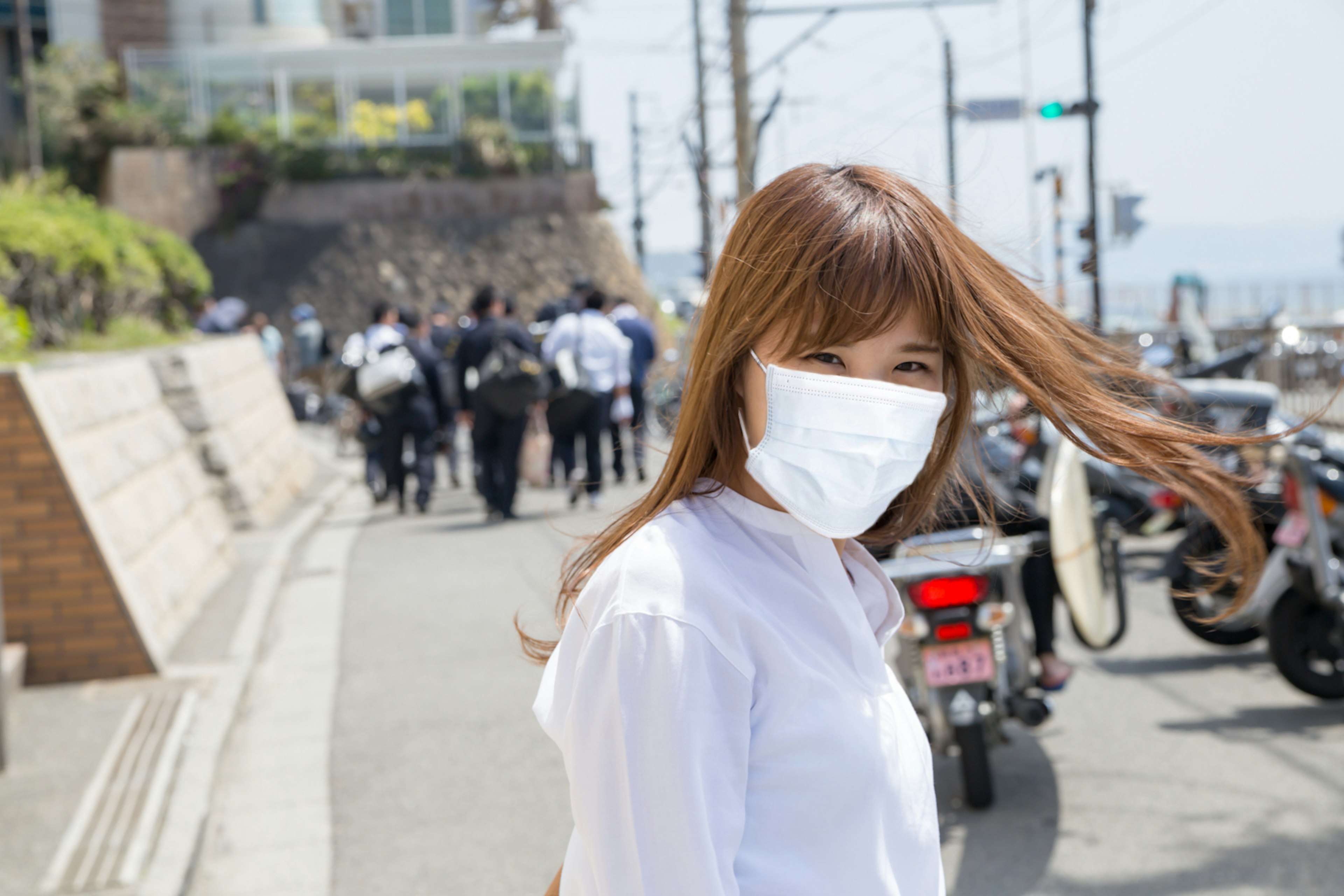 Seorang wanita mengenakan kemeja putih dan masker berjalan di jalan dengan orang-orang dan sepeda motor di latar belakang