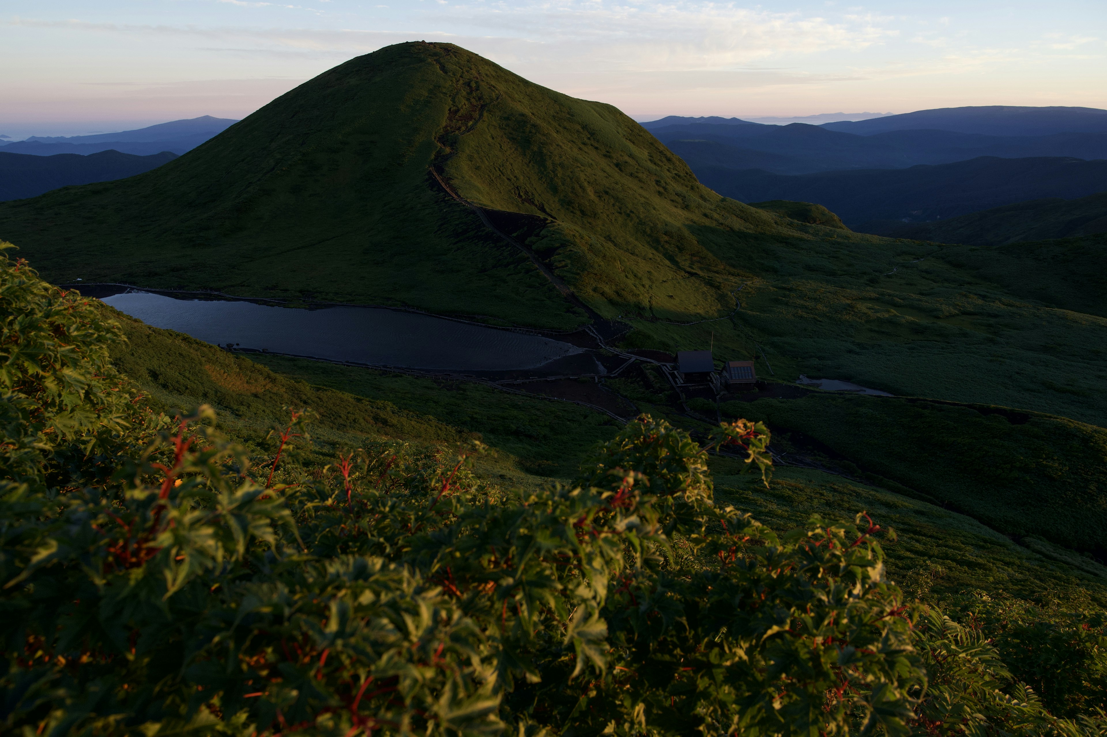 Bukit hijau dengan danau tenang di latar depan yang diterangi cahaya matahari terbenam