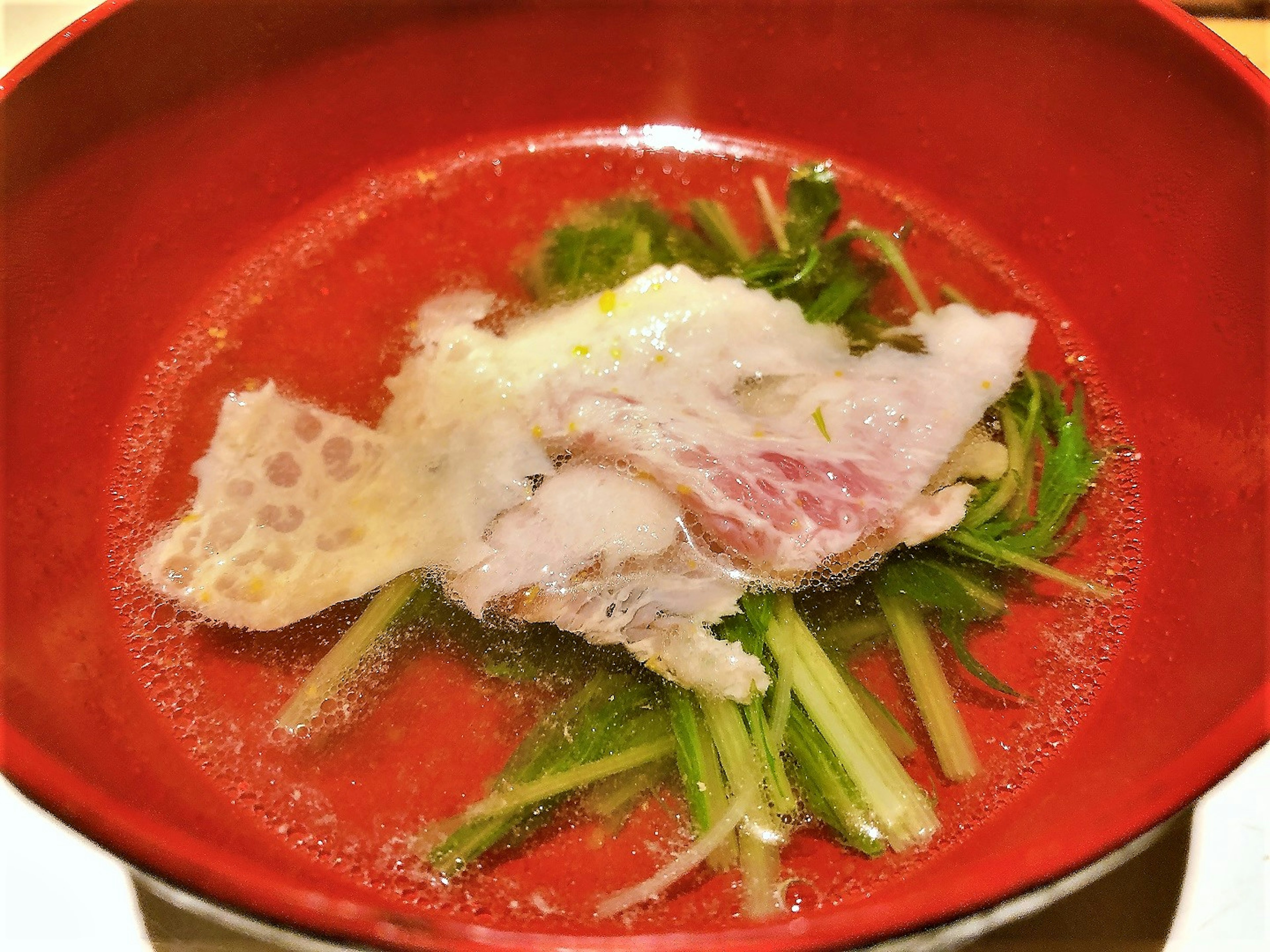 A red bowl containing soup with fresh fish slices and green vegetables